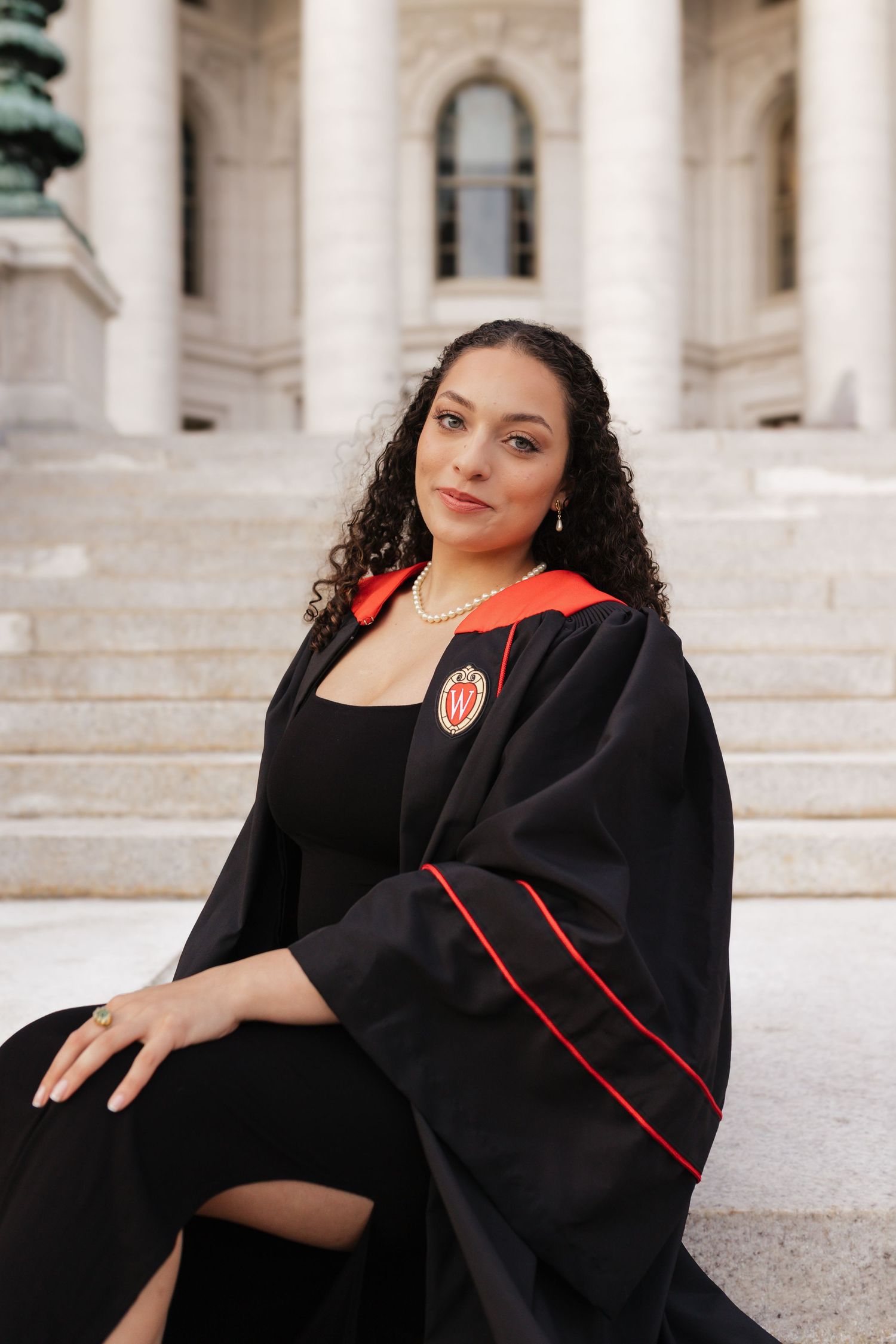 Graduate wearing academic regalia with red trim poses on steps of classical building.