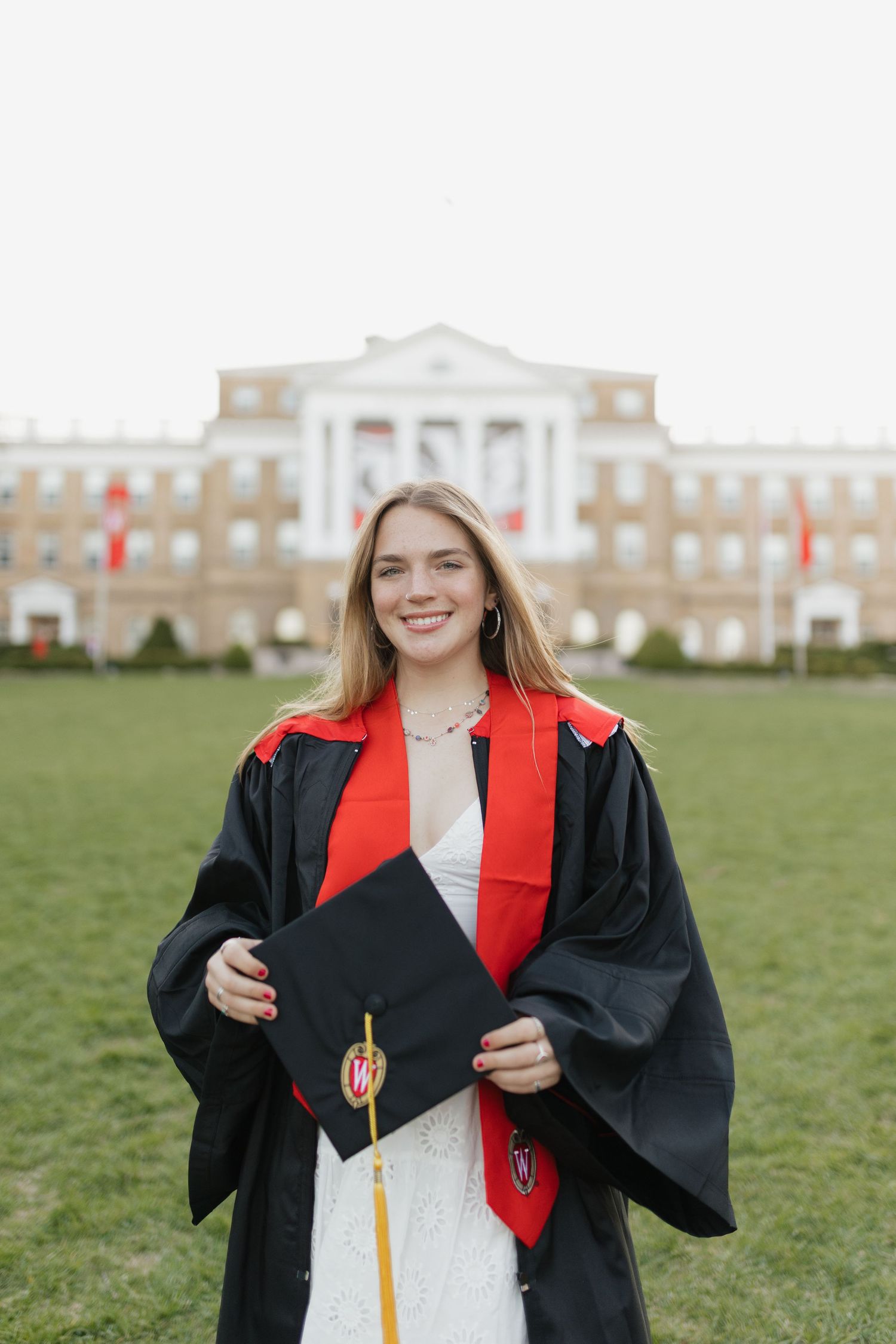 Graduate in black gown and red stole poses on grass with academic building in background.