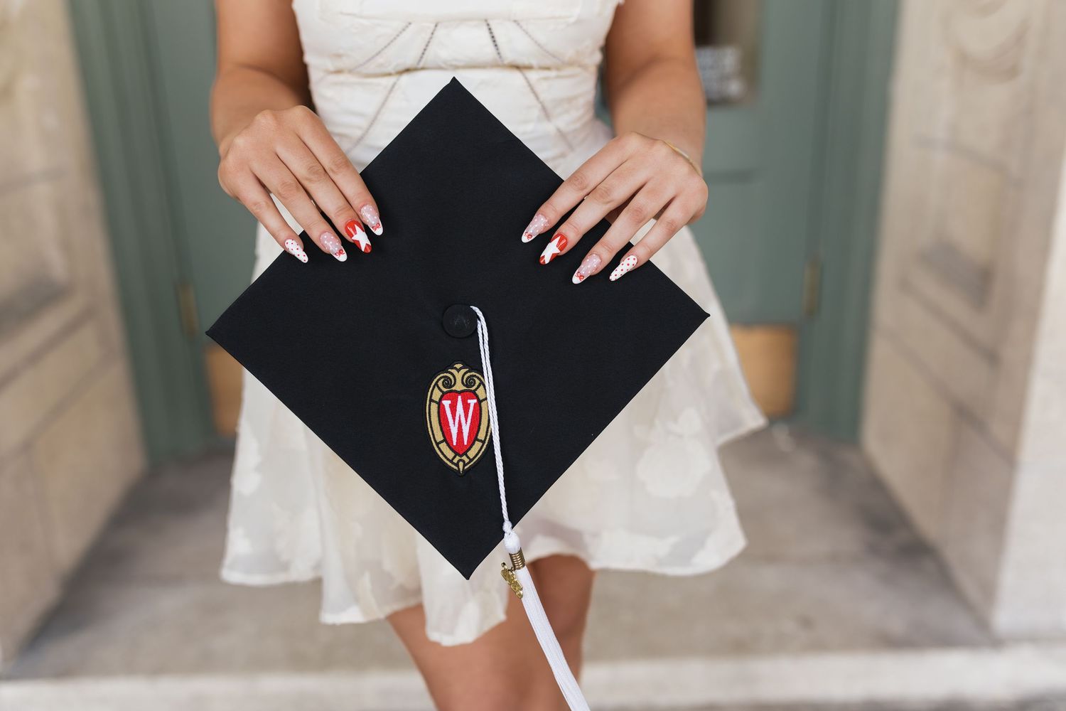 Graduates hold black mortarboards while wearing white dresses in an elegant hallway.