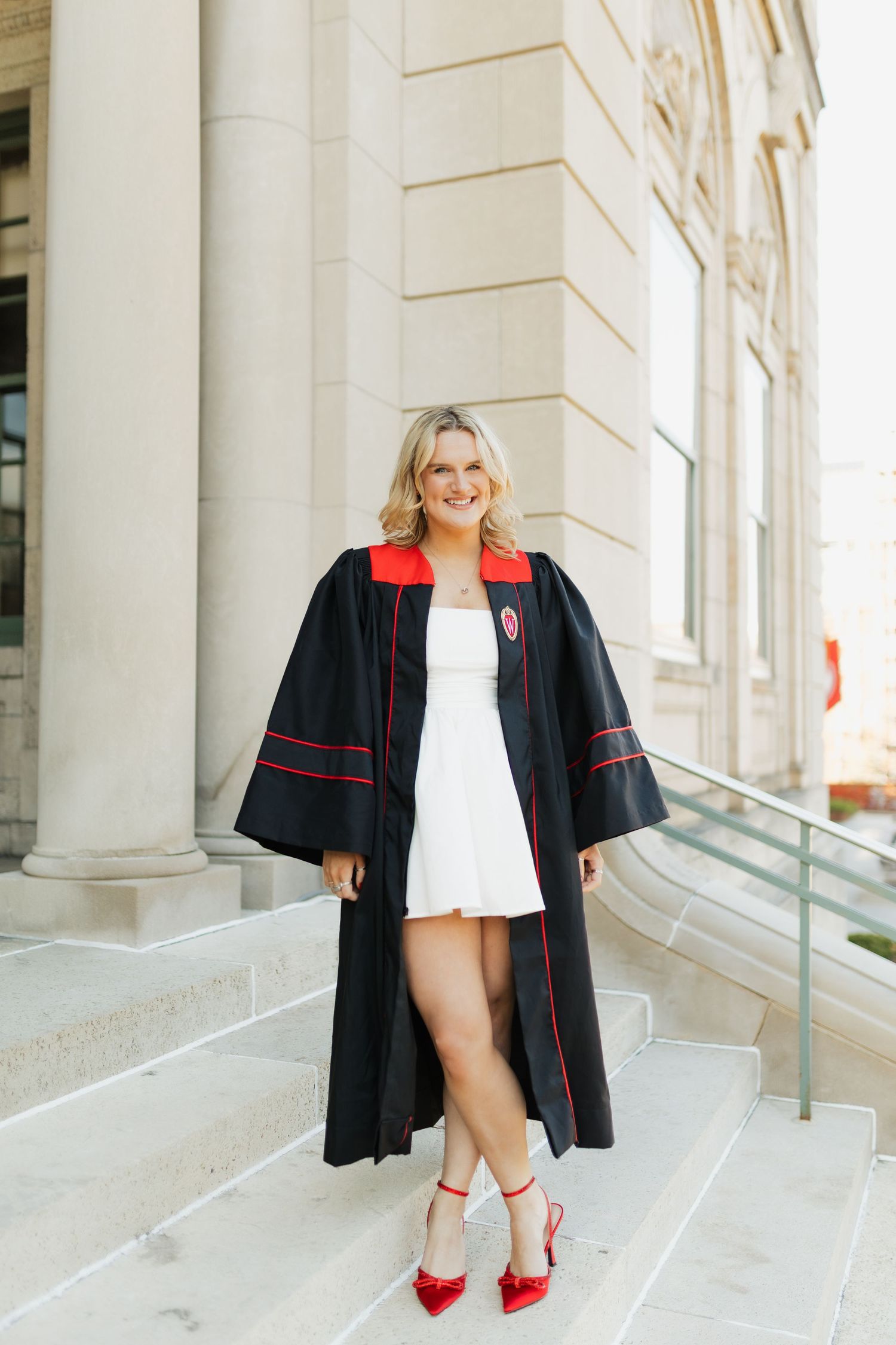 Graduate in black gown with red trim posing along white columned walkway.