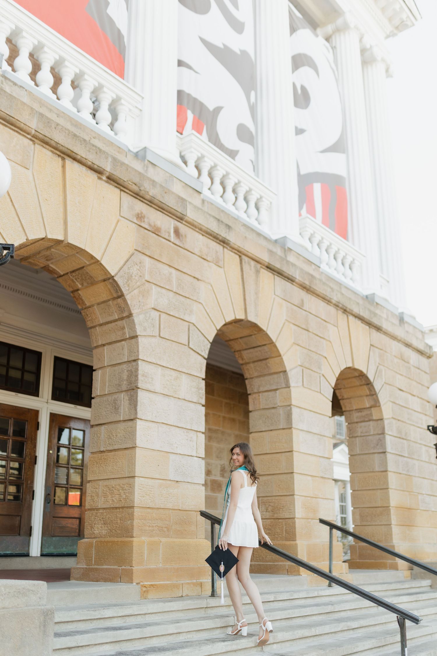 Graduate in white dress stands on stone steps of classical university building with arched entryways.