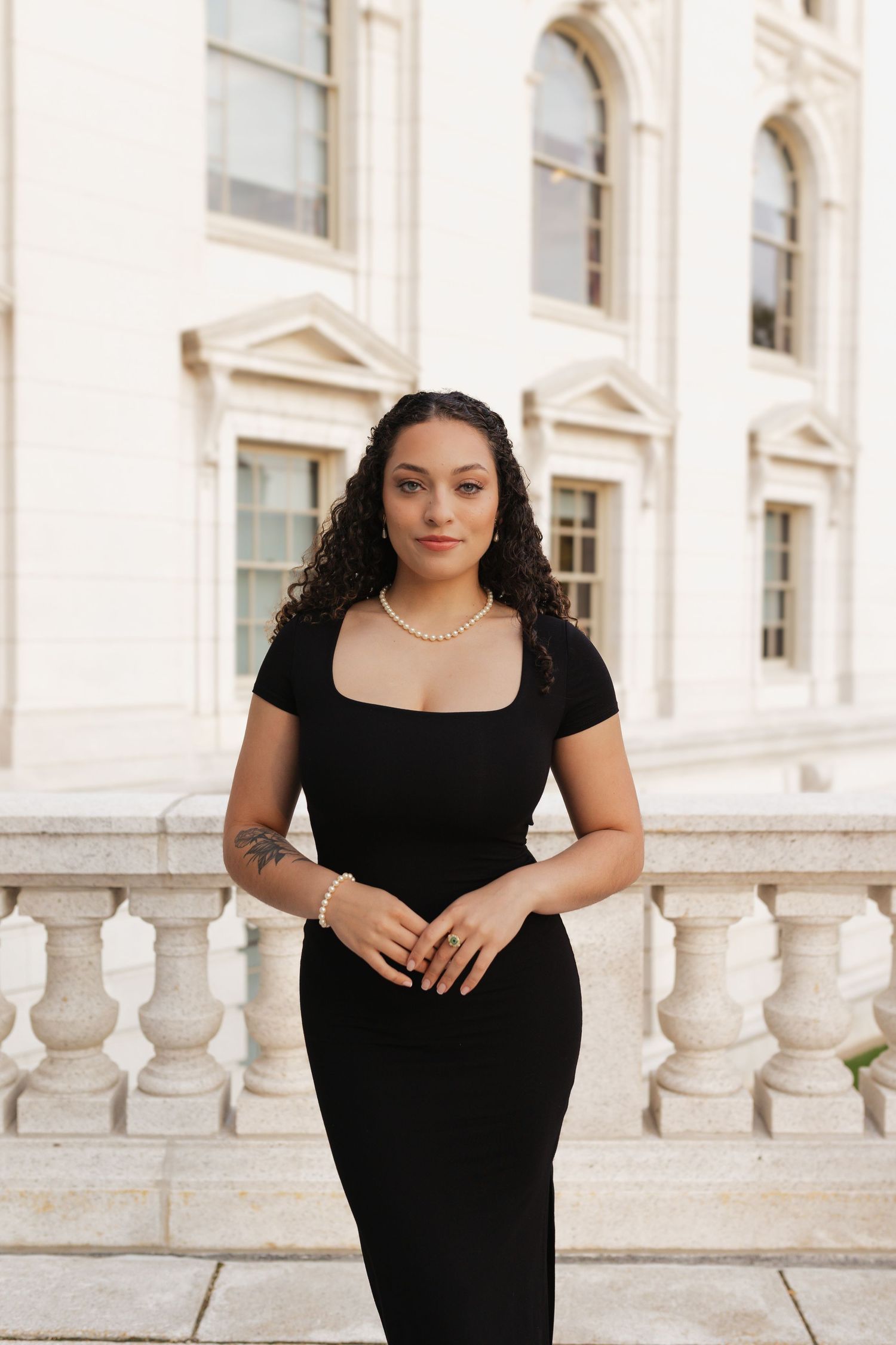 Graduate in black dress poses against white stone balustrade of classical building.