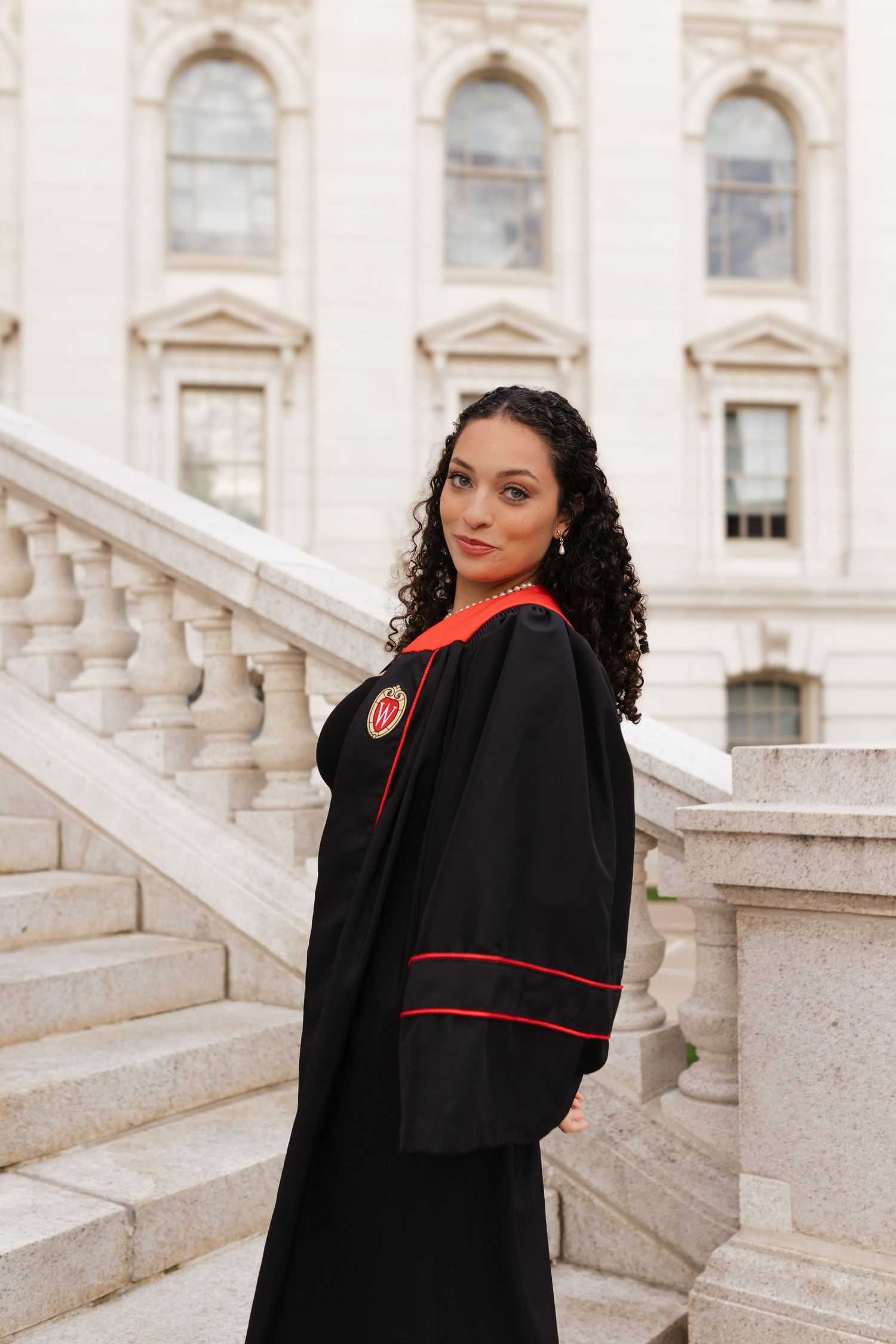 Graduate in black graduation gown with red trim poses on grand stone staircase.