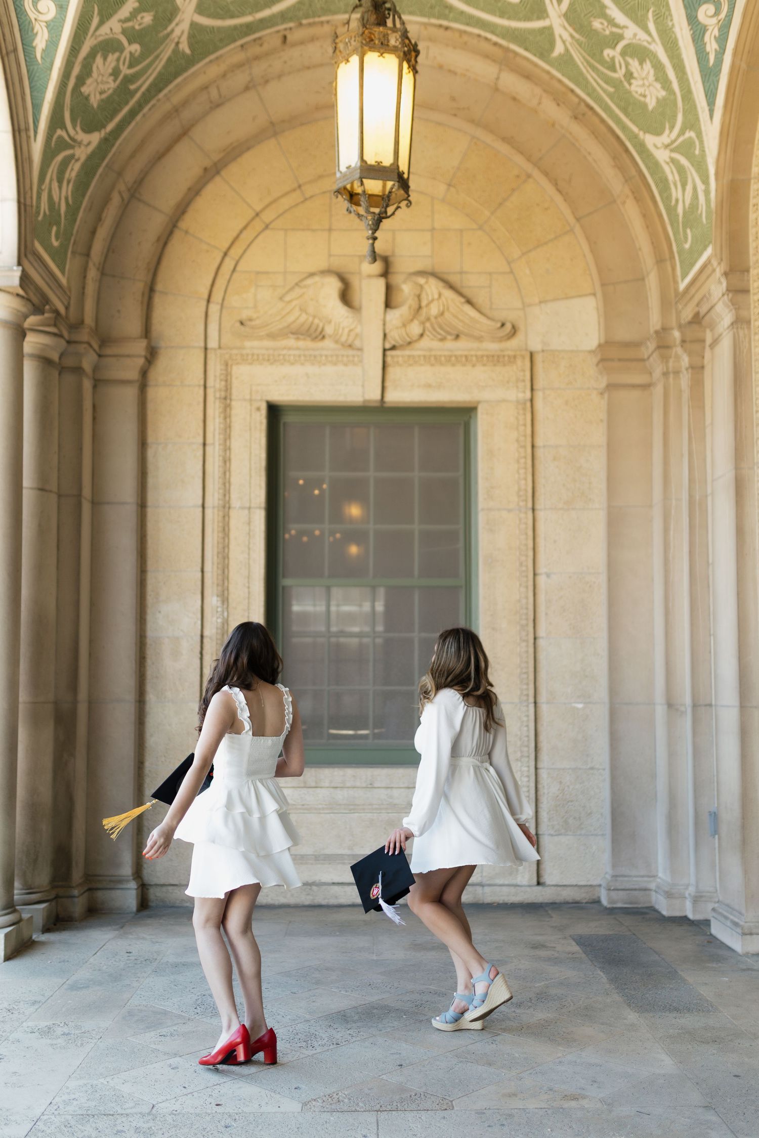 Graduates in white dresses pose playfully in an ornate stone archway on campus.