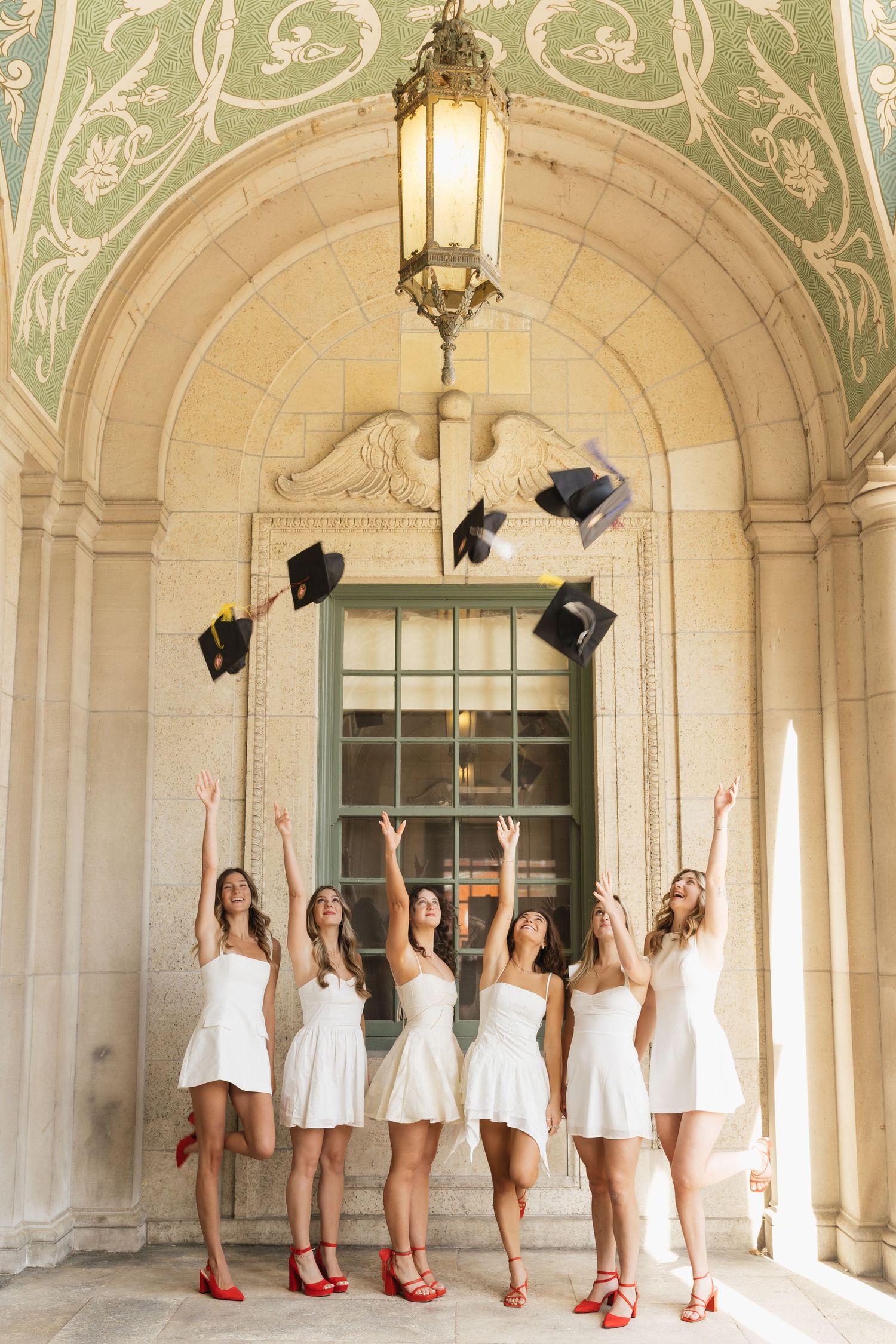 Five graduates in white dresses throw their caps under an ornate archway.
