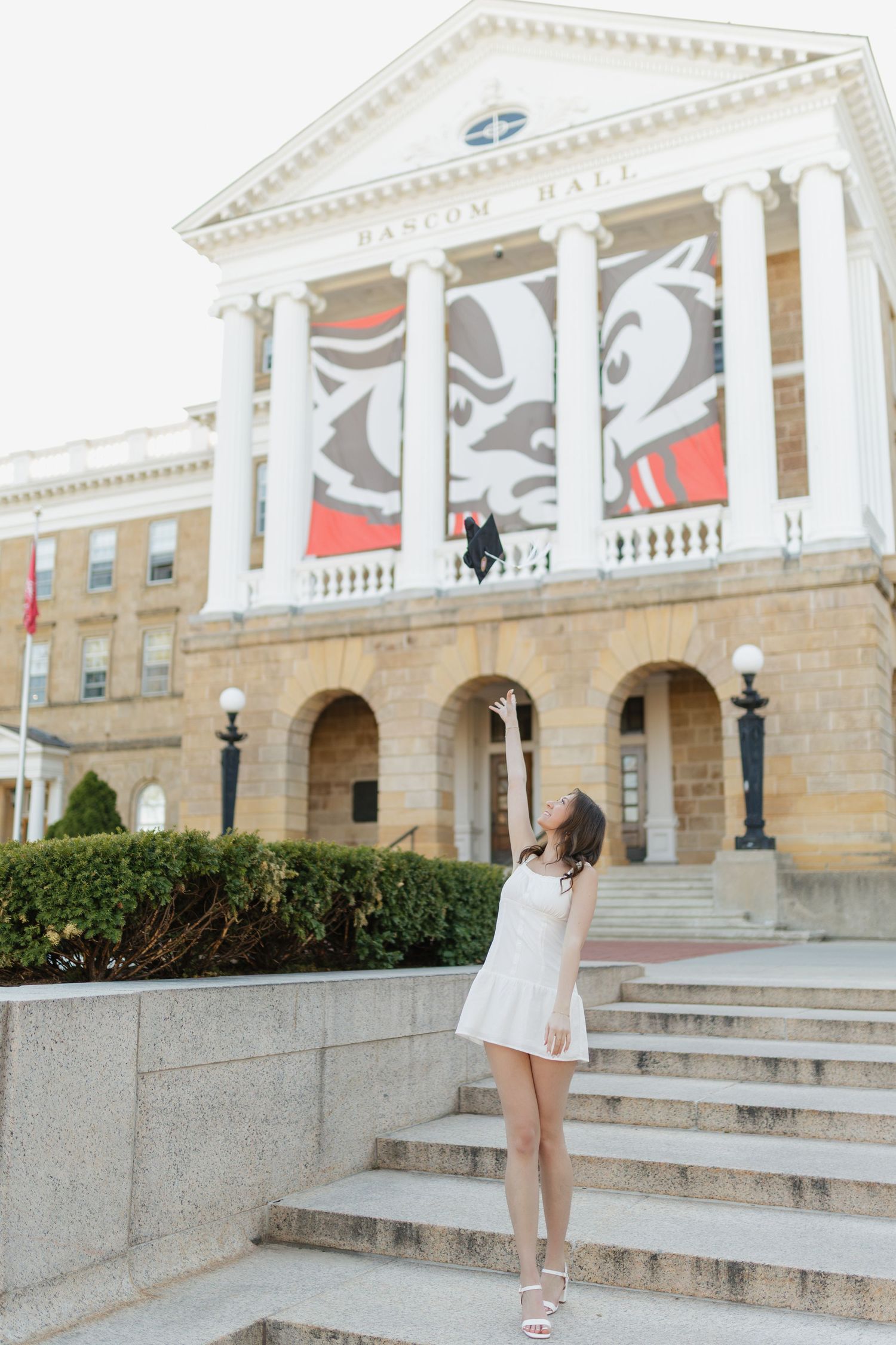 Graduate in white dress poses on steps of grand university building with decorative columns and facade.