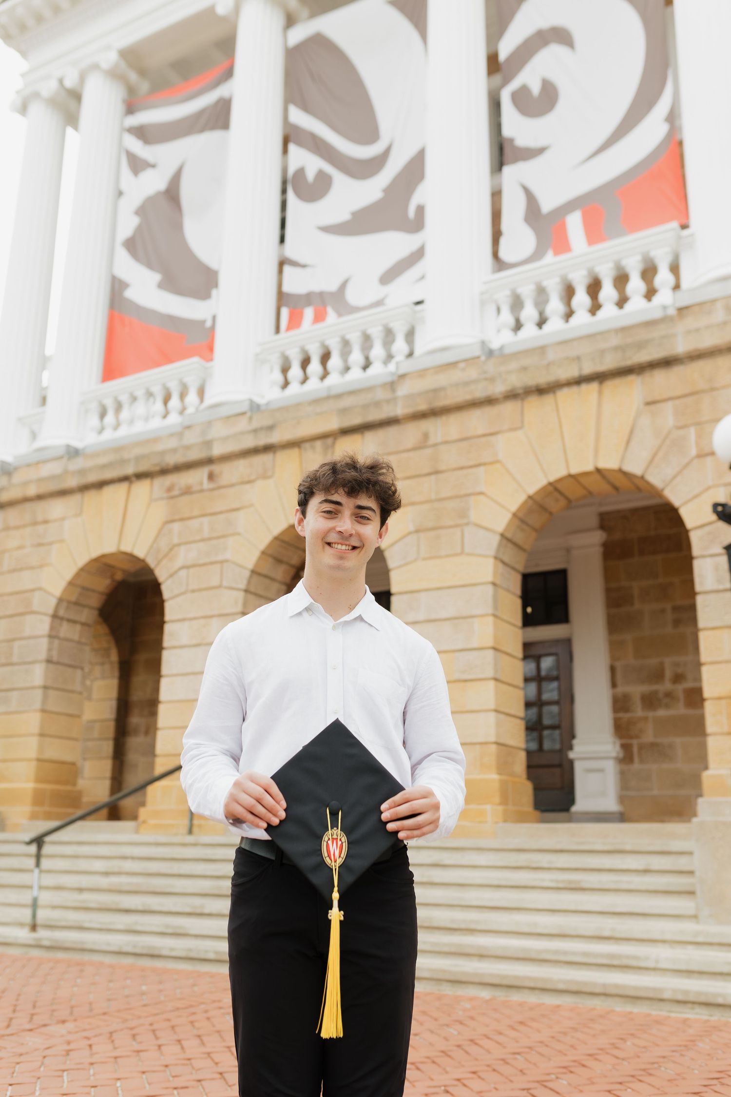 Graduate holding graduation cap with gold tassel against decorative university building backdrop.