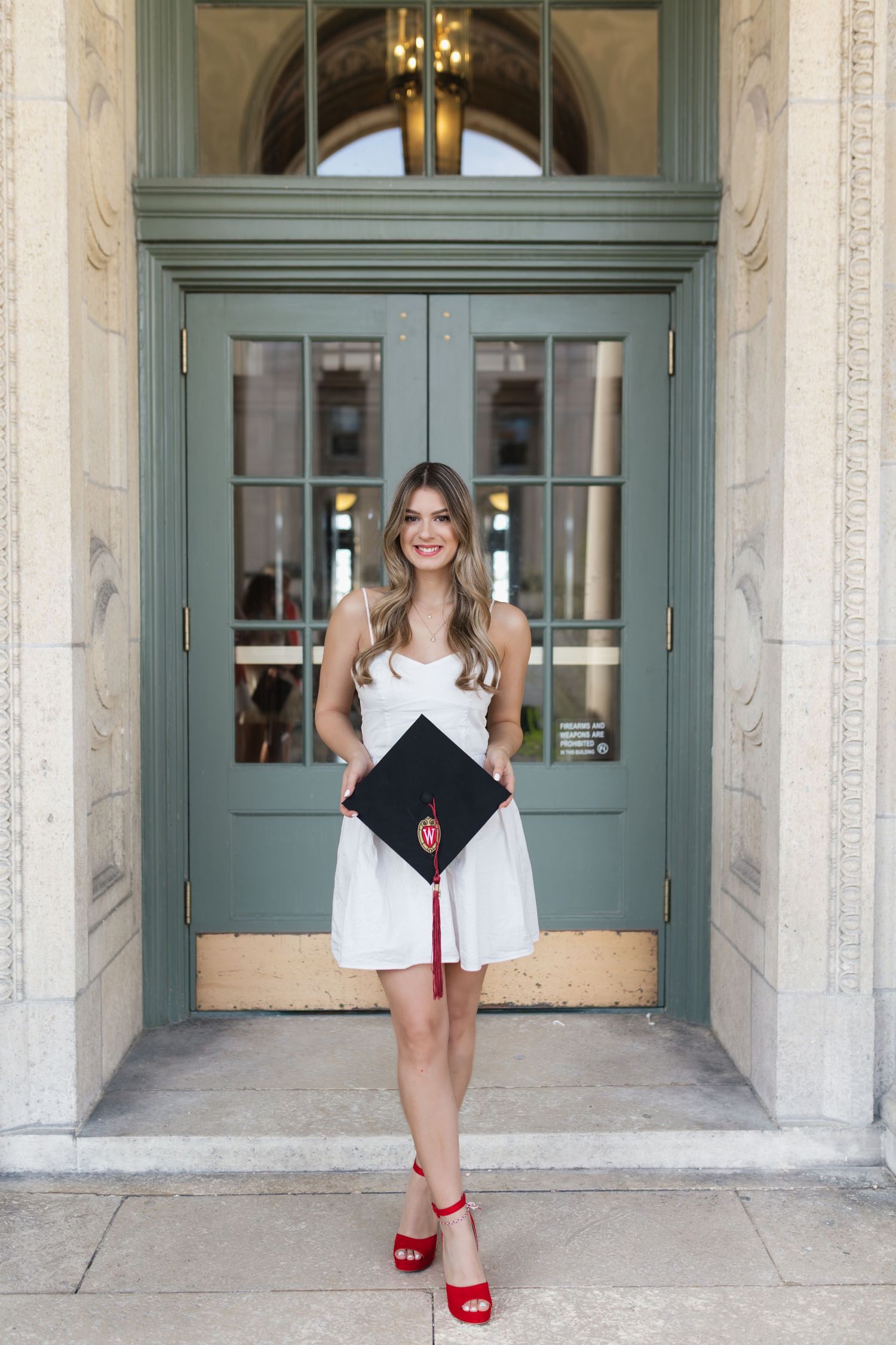 Graduate poses playfully with mortarboard in elegant stone hallway.