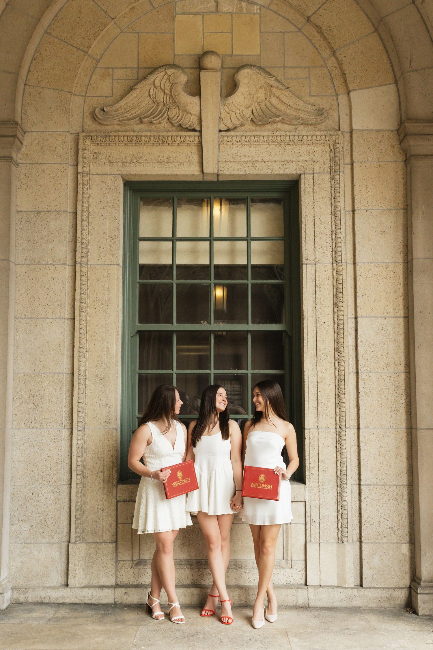 Three graduates in white dresses pose in front of an ornate stone window with red diplomas.
