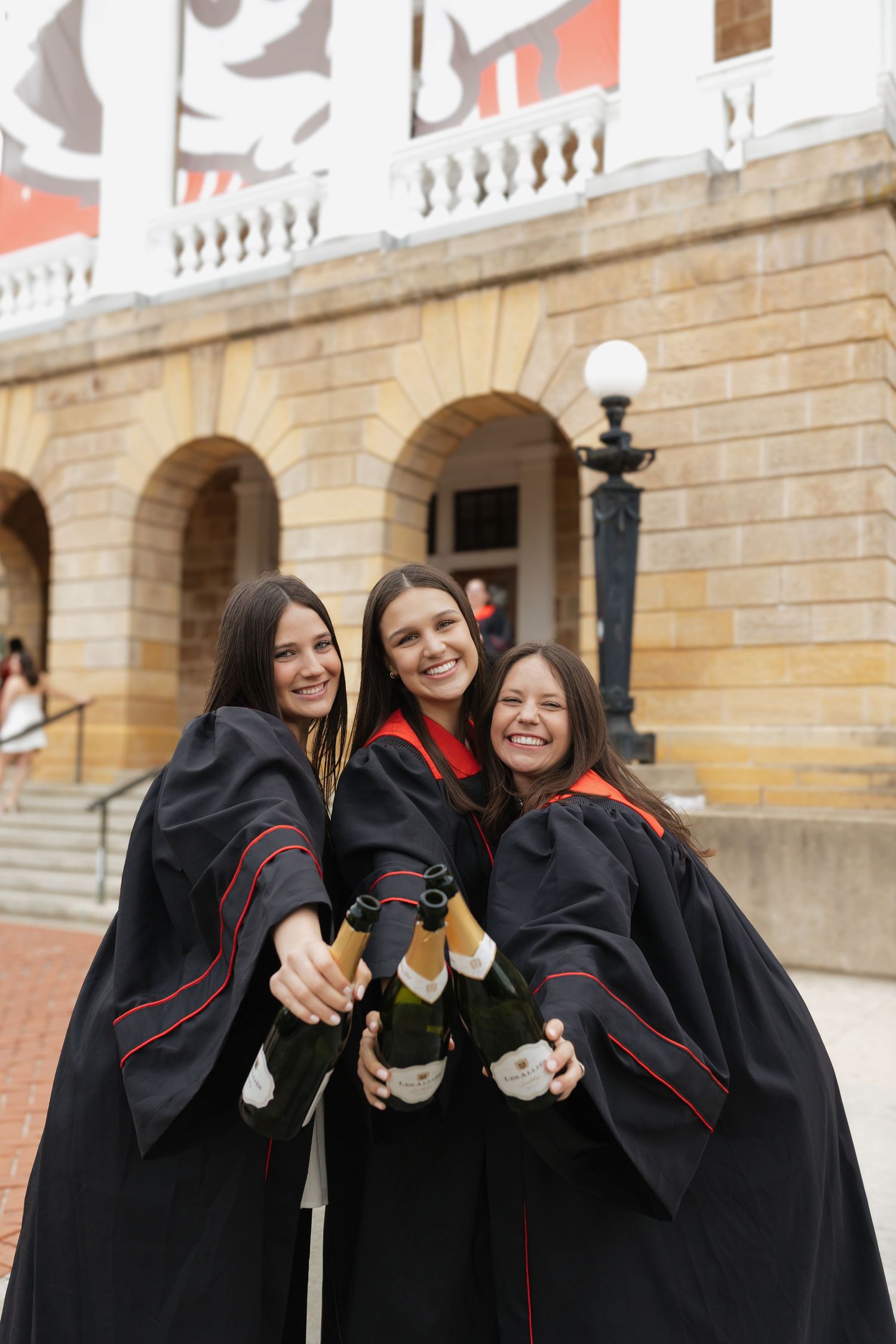 Three graduates in black graduation gowns celebrate with champagne bottles outside a historic university building.