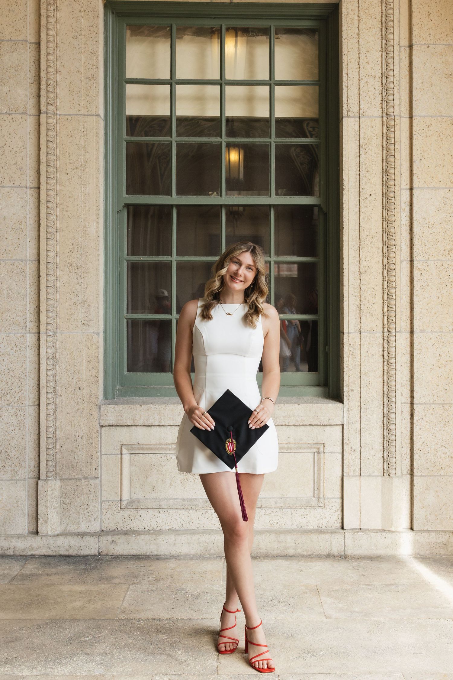 Graduate poses in white dress and red shoes under stone archway.