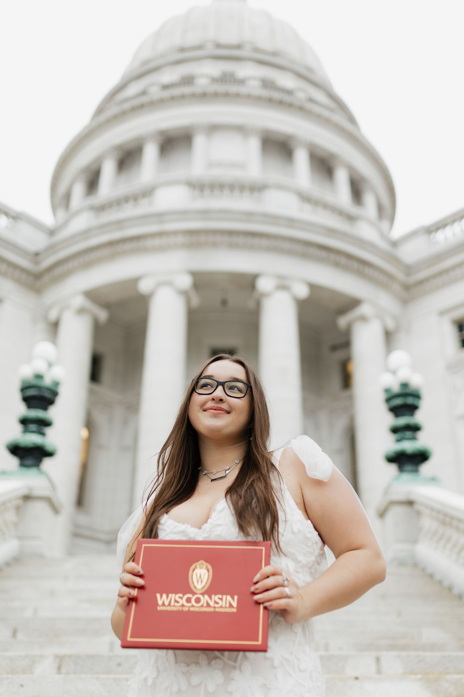 Graduate holds red Wisconsin diploma folder in front of domed capitol building.