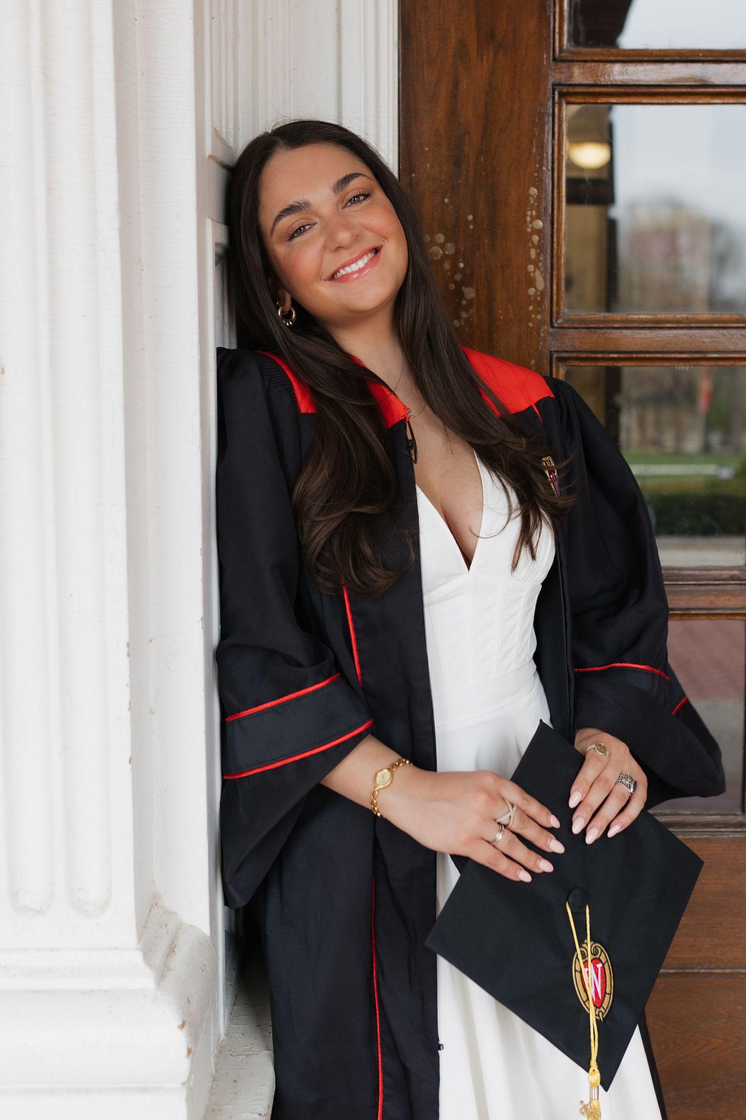 Graduate posing in black graduation gown with red trim against white columns.