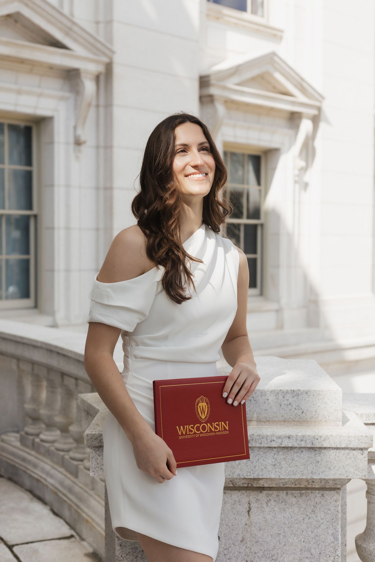 A UW Madison graduate in a white dress poses with their diploma on stone steps of the Wisconsin State Capitol Building.