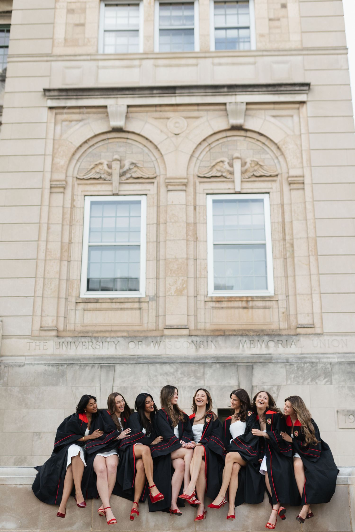 Group of graduates in black robes sit together on stone steps of campus building.