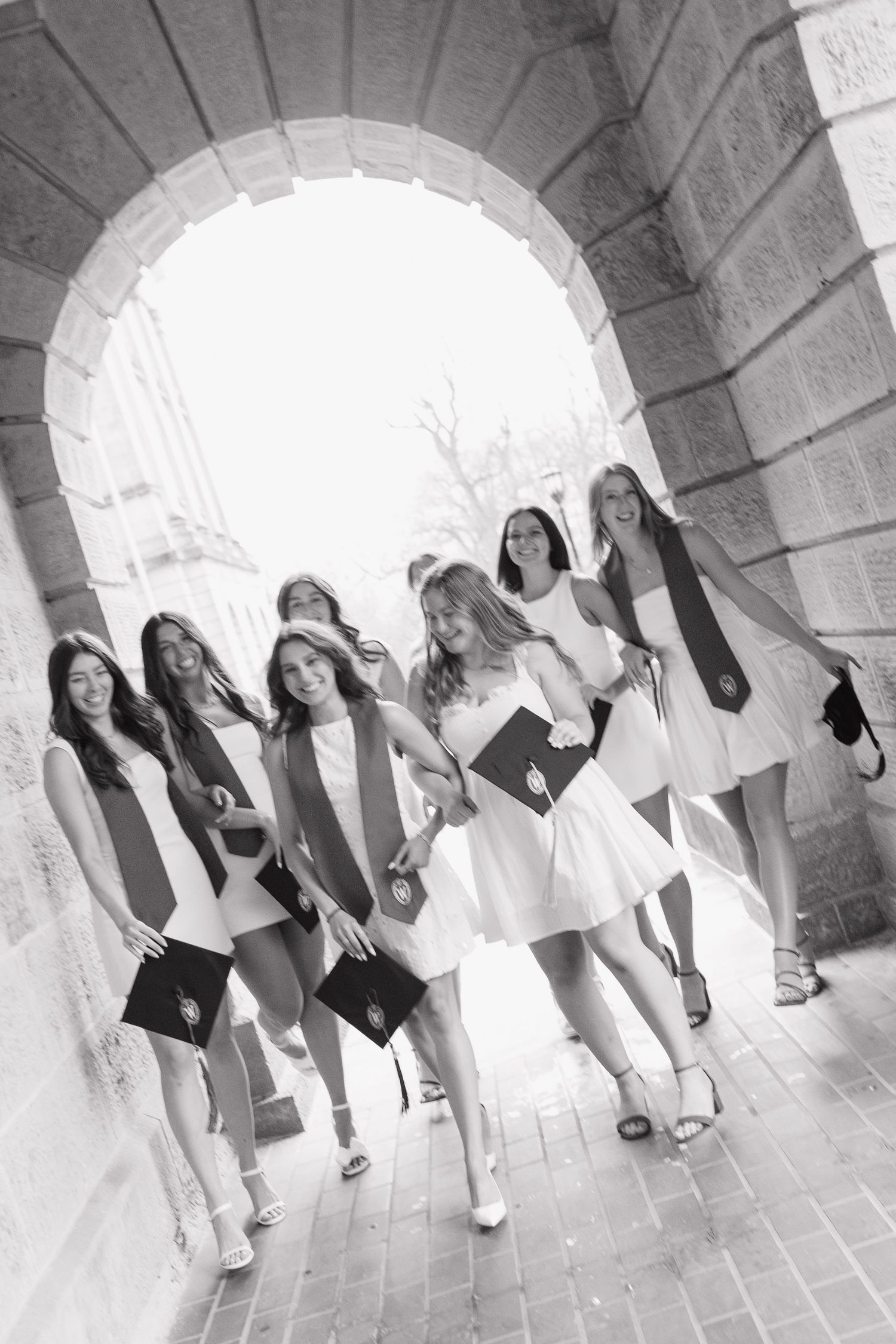 Group of graduates in white dresses pose under stone archways.
