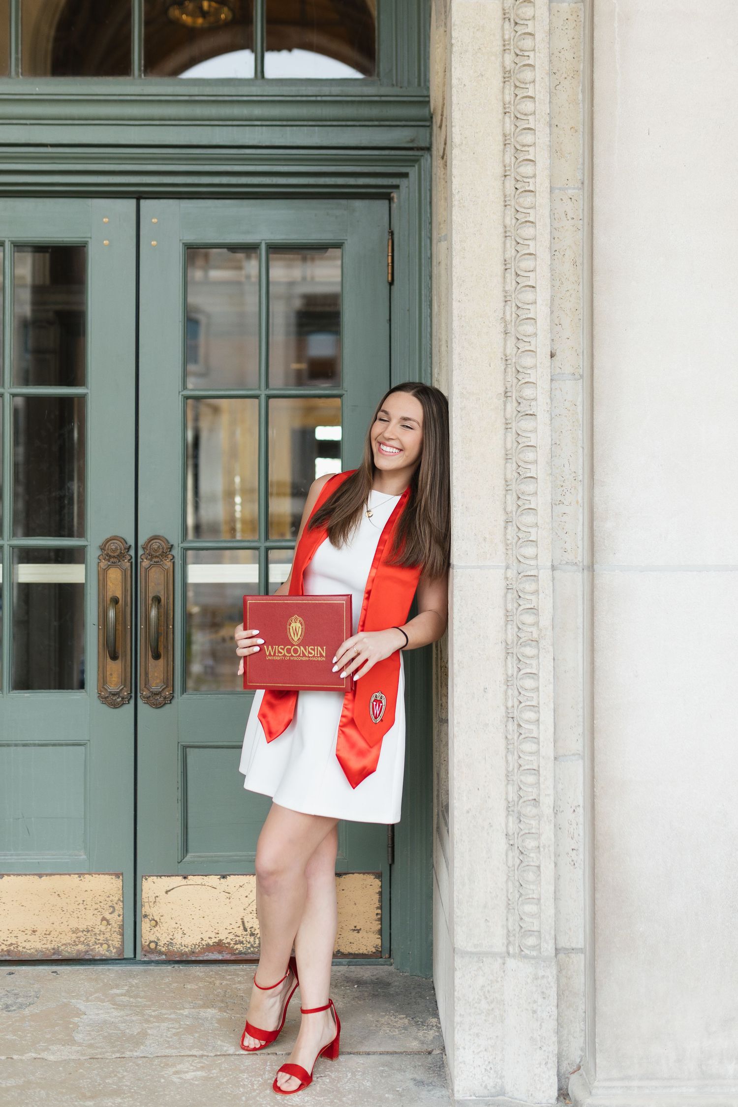 Graduate in white dress and red accessories poses by vintage green door.
