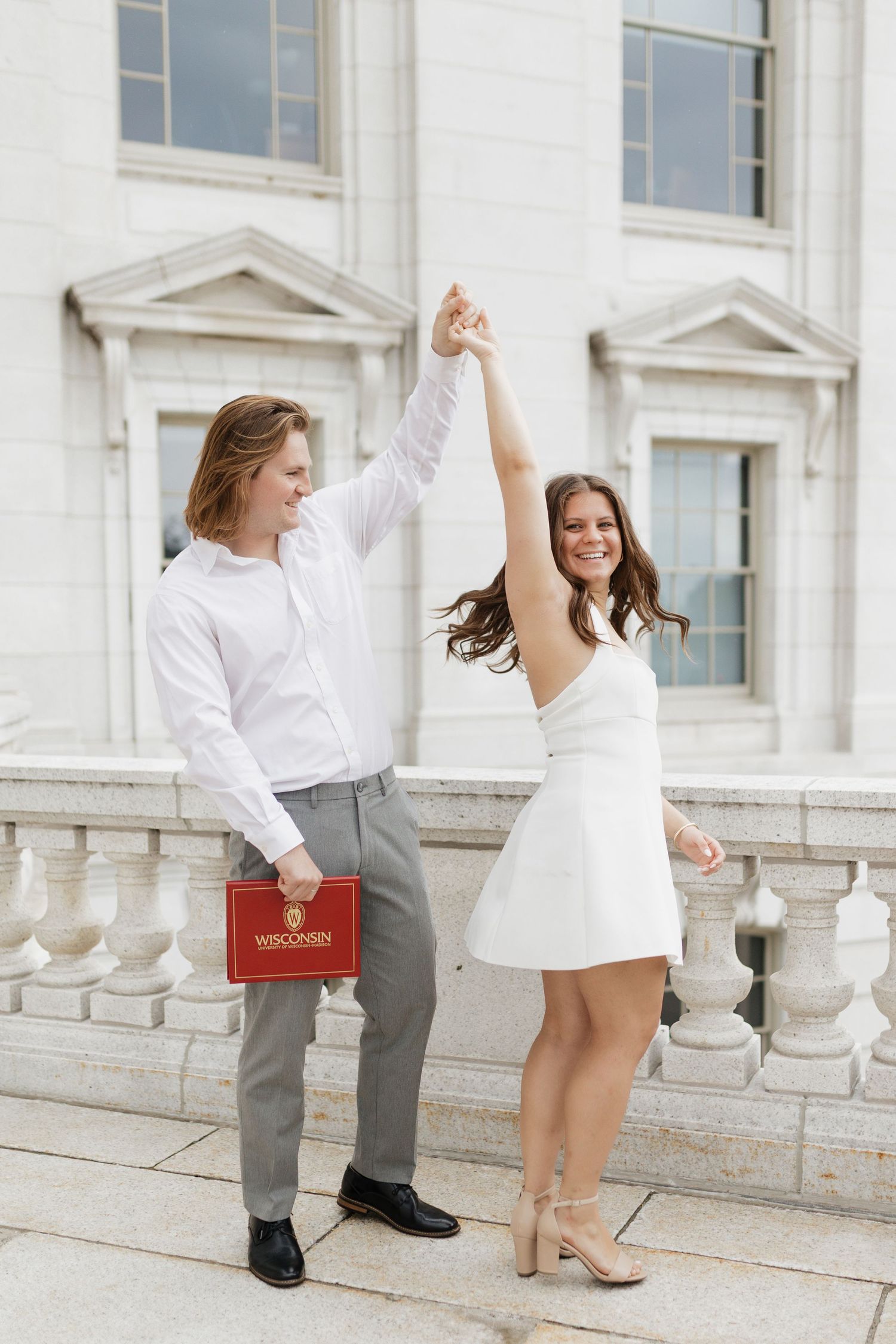 Graduates in formal attire pose playfully on white stone steps of a university building.