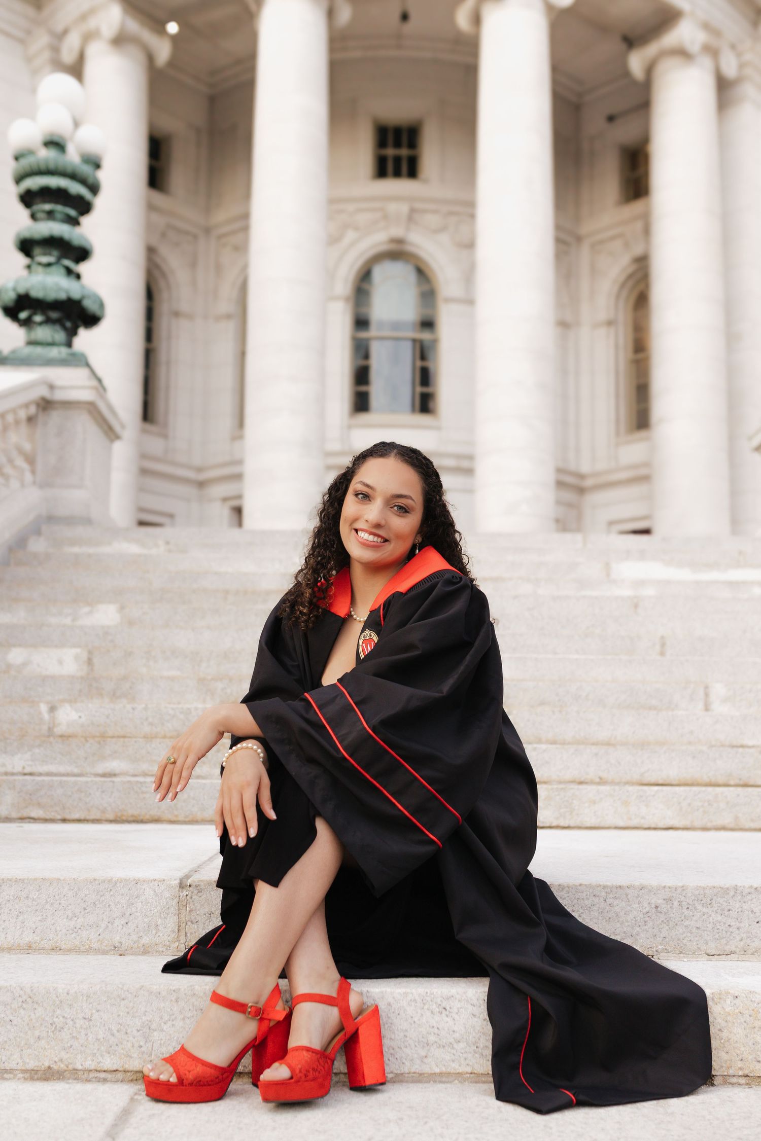Graduate in academic regalia and red heels poses playfully on marble steps.