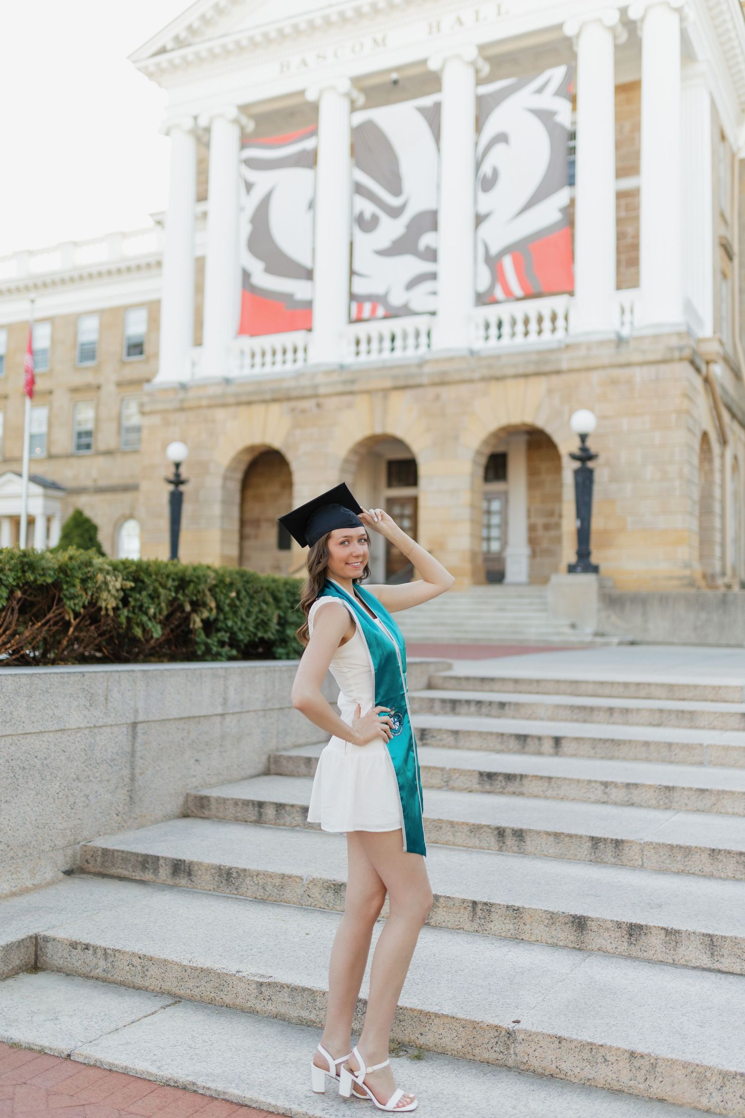 Graduate in white dress and teal sash poses on university steps with decorative building facade.