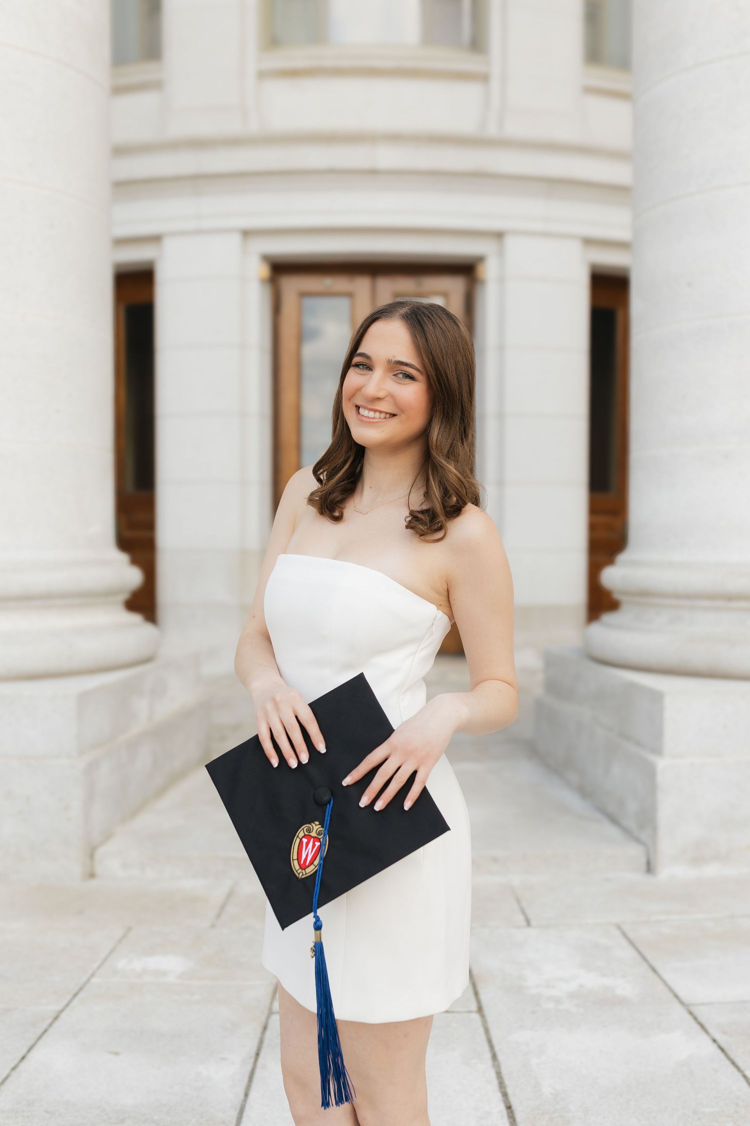 A UW Madison graduate in a white dress poses with their graduation cap at the Wisconsin State Capitol Building.