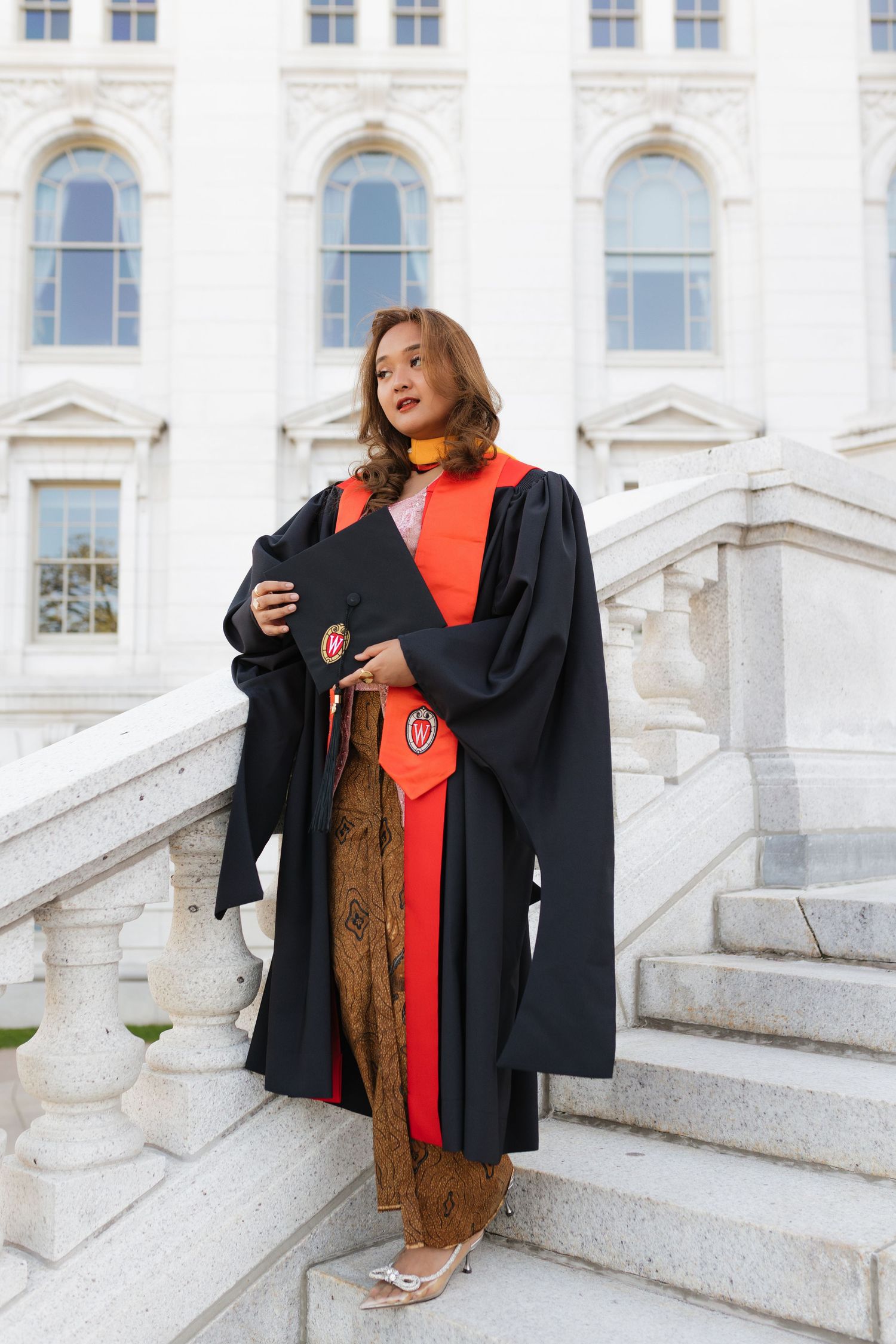 Graduate in academic regalia posing on white marble steps of historic building.