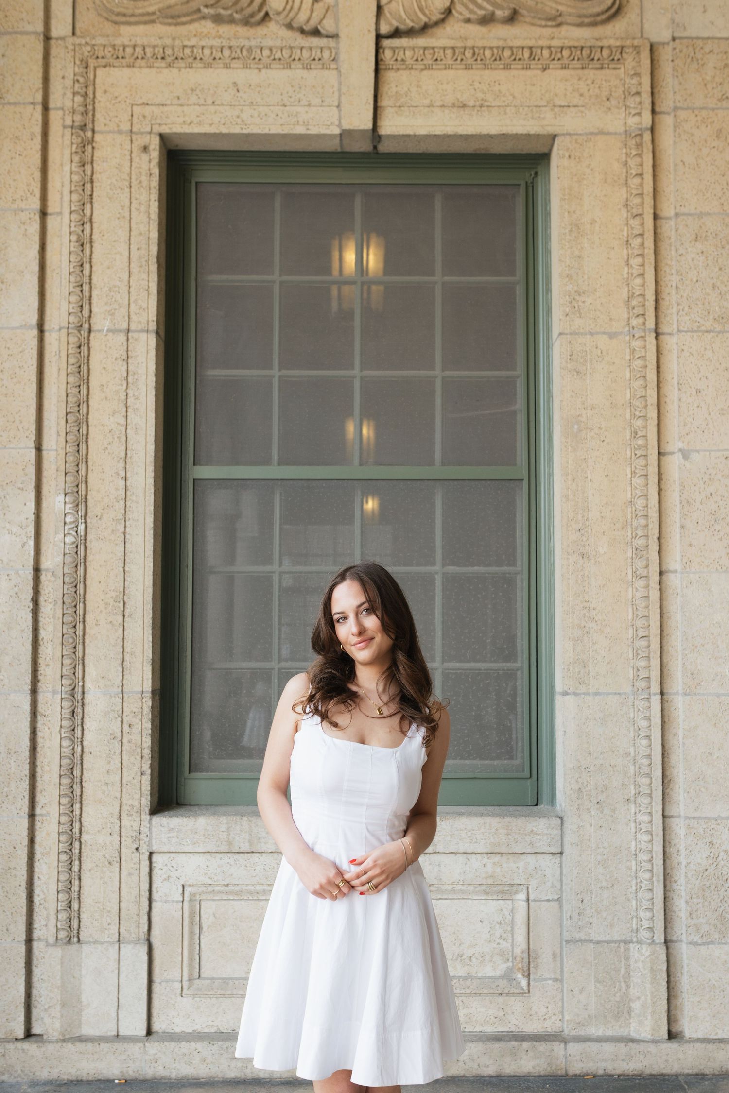 Graduate in white dress poses elegantly against a stone building with green window frame.