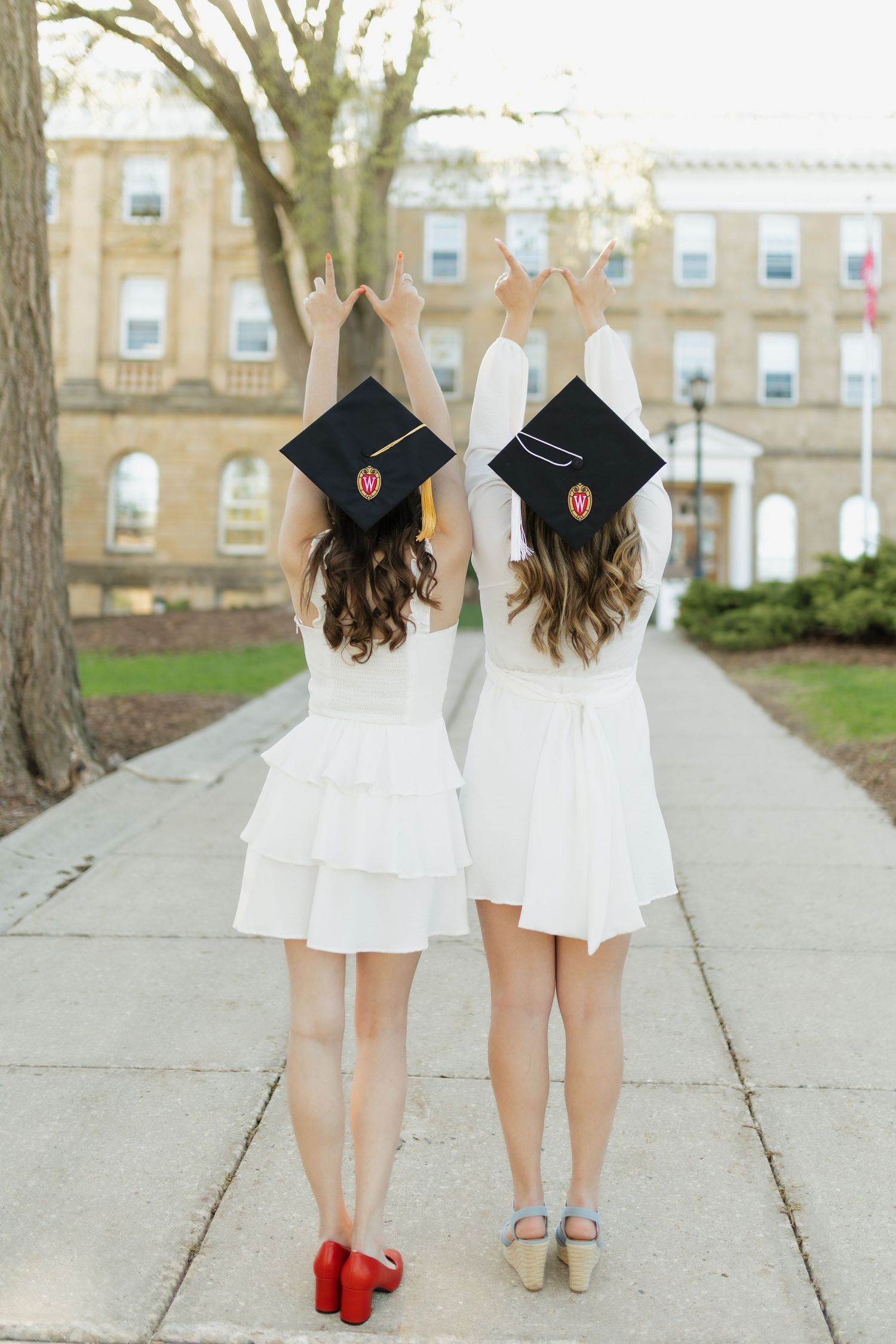 Two graduates in white dresses and black caps walk together showing their graduation shoes.