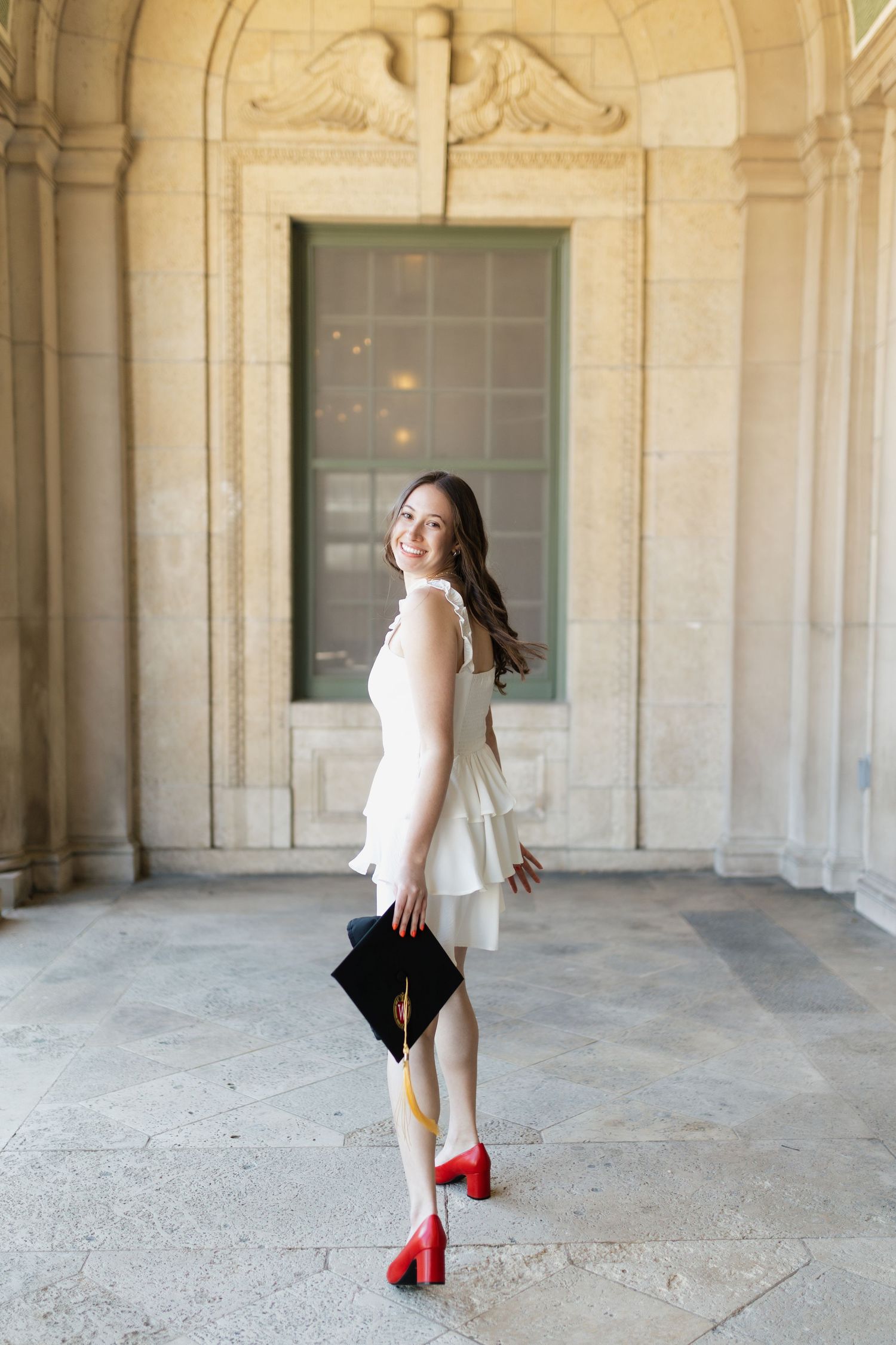Graduate in white dress and red heels poses playfully in stone archway corridor.
