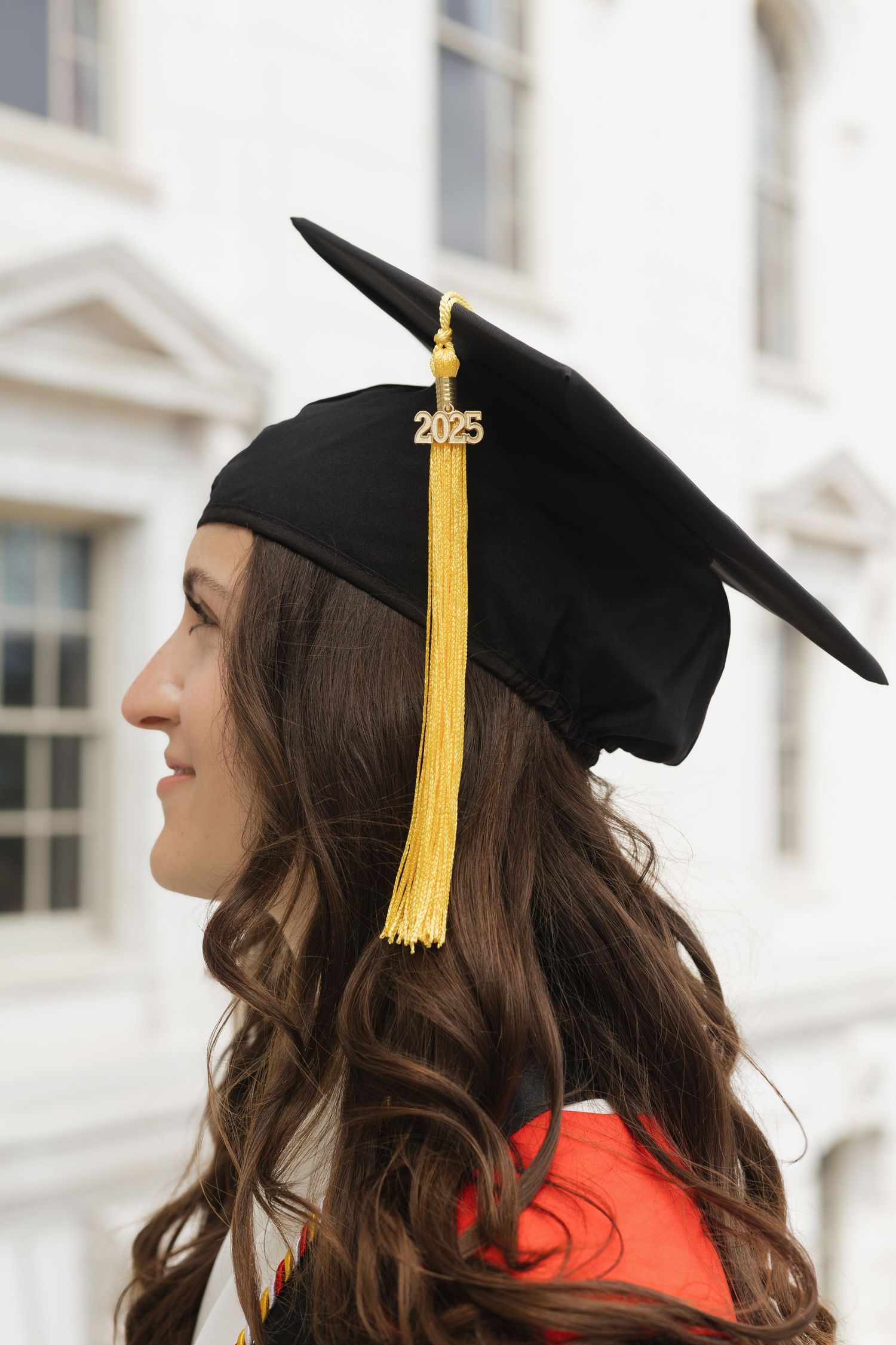 Graduate in cap with gold tassel poses in profile against white building.