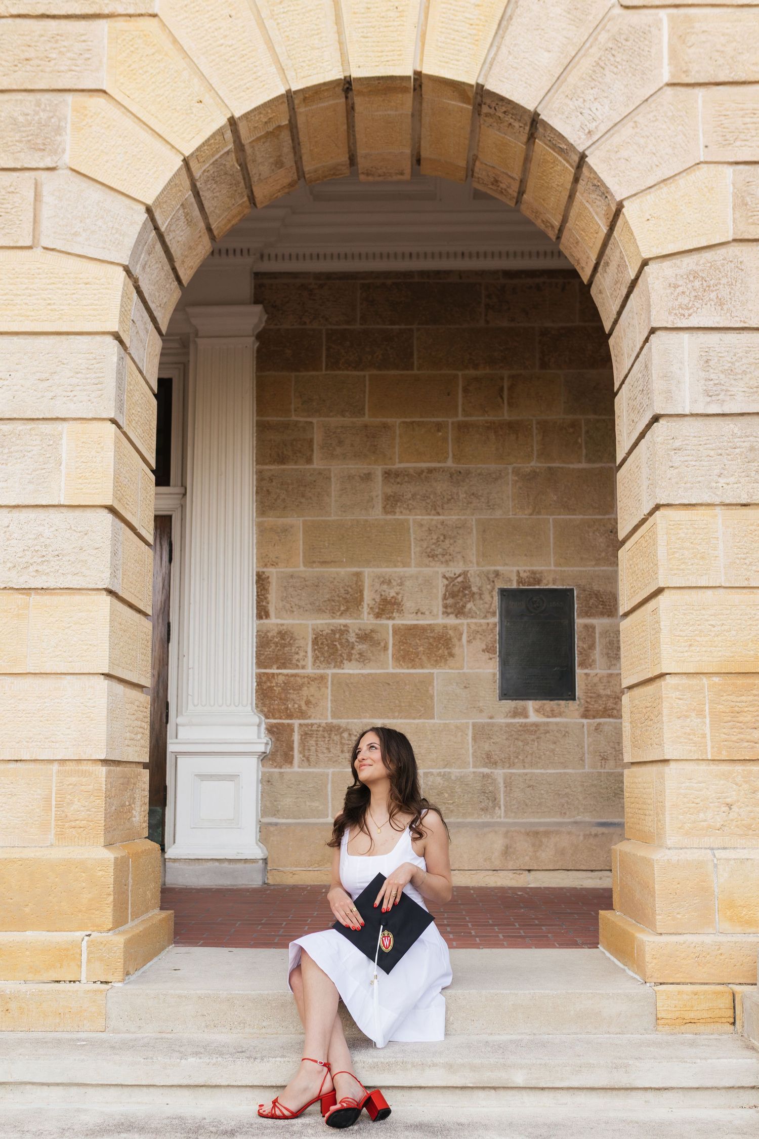 A graduate in red heels poses under stone archways of a university building.