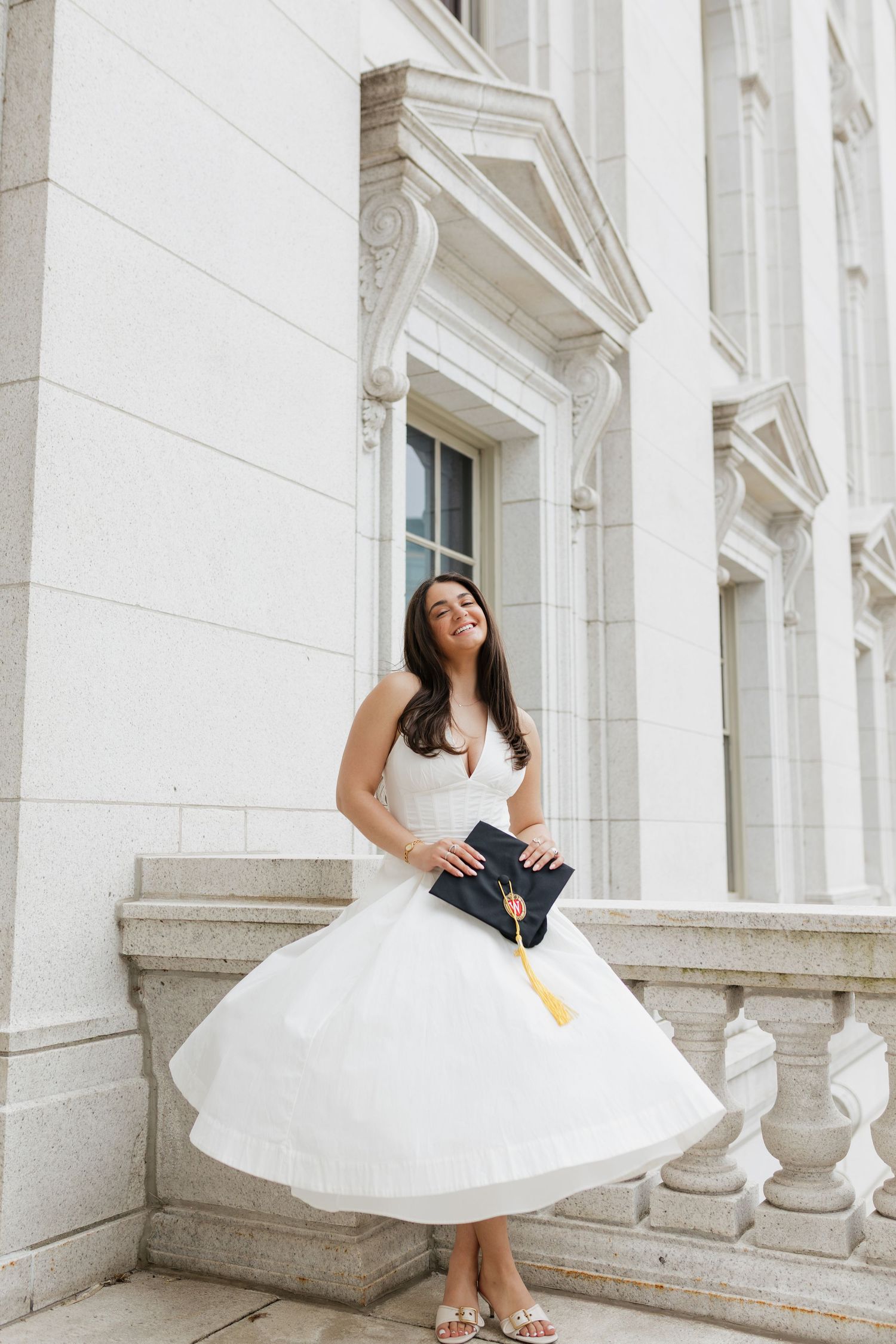 A UW Madison graduate in a white dress poses with their graduation cap at the Wisconsin State Capitol Building.