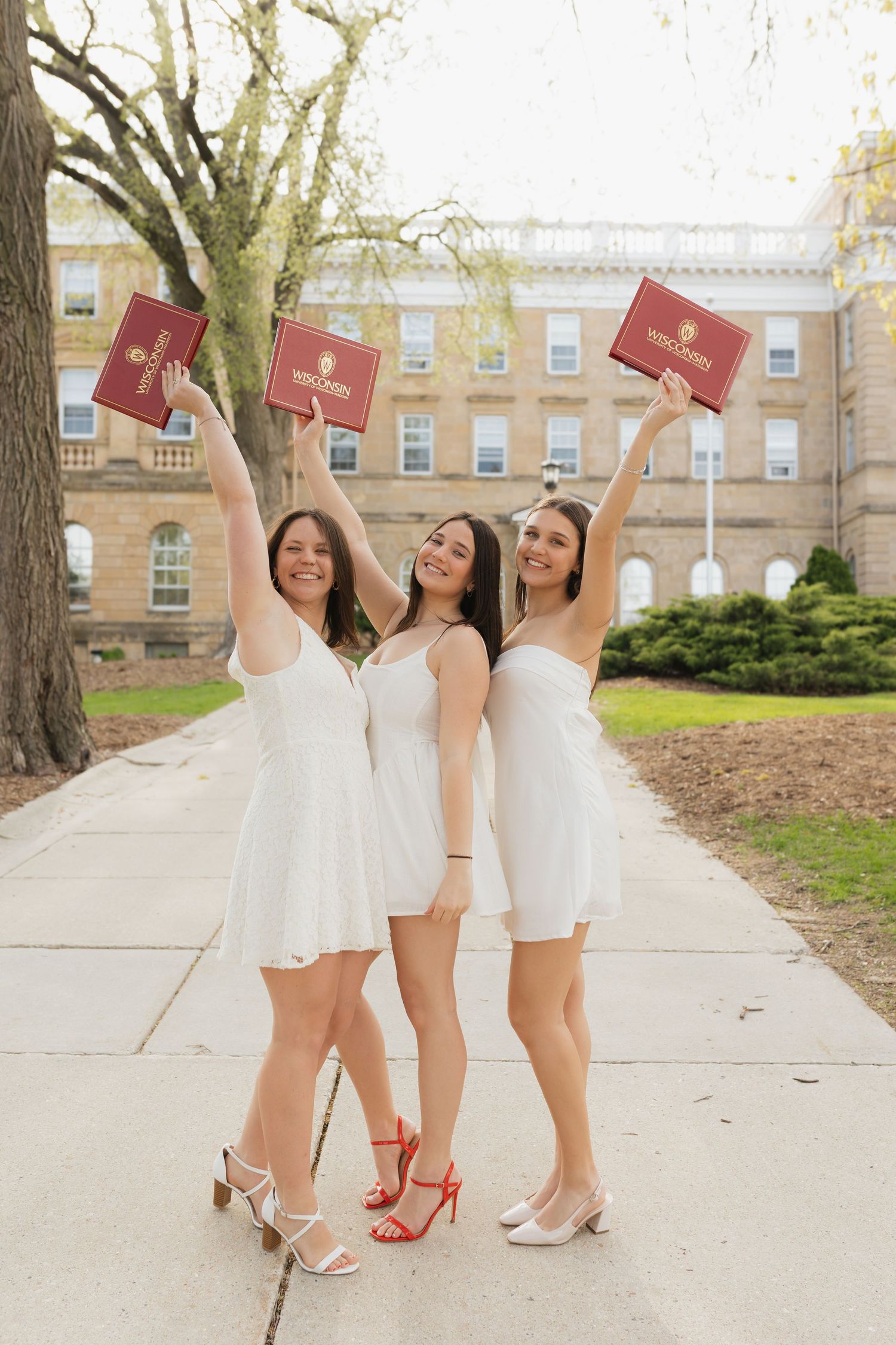 Three graduates in white dresses hold up diplomas while showing off their shoes.