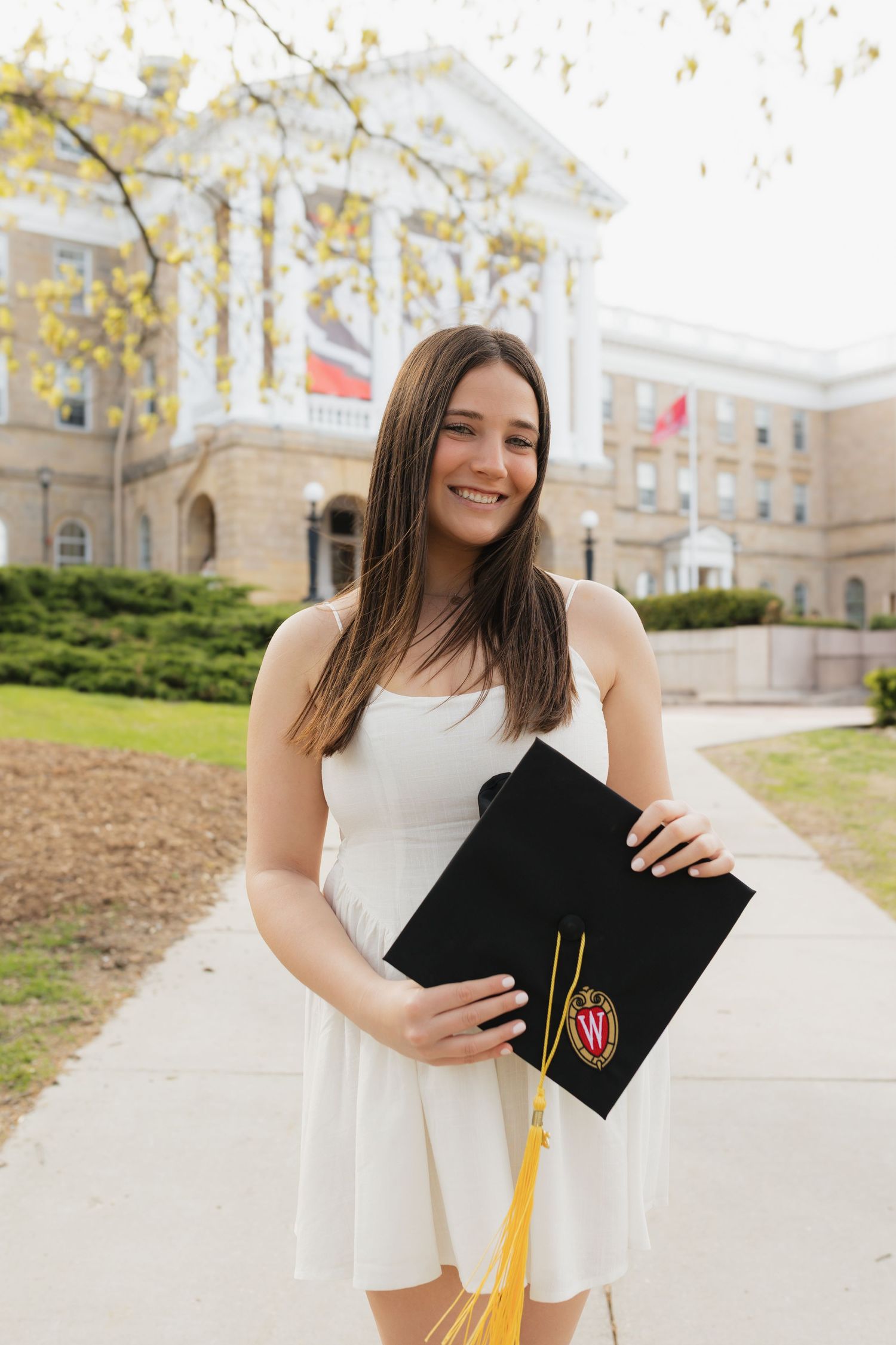 Graduate in white dress holds graduation cap with gold tassel on campus grounds.