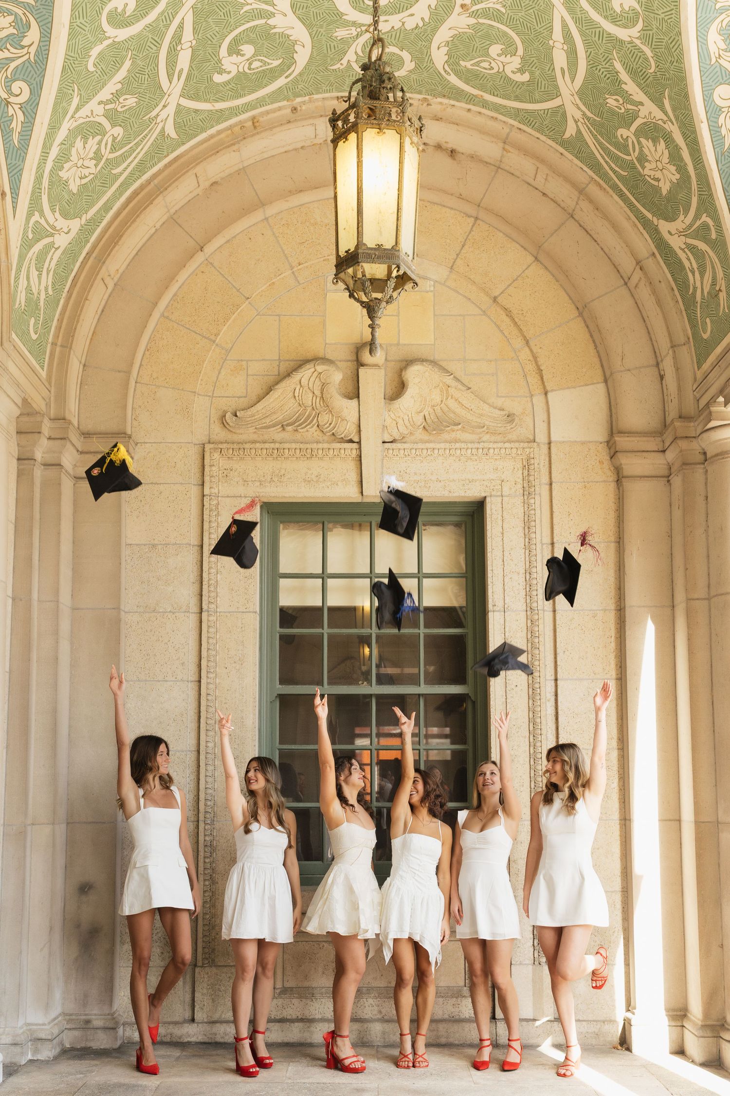 Graduates in white dresses throw caps in an ornate hallway with decorative ceiling.