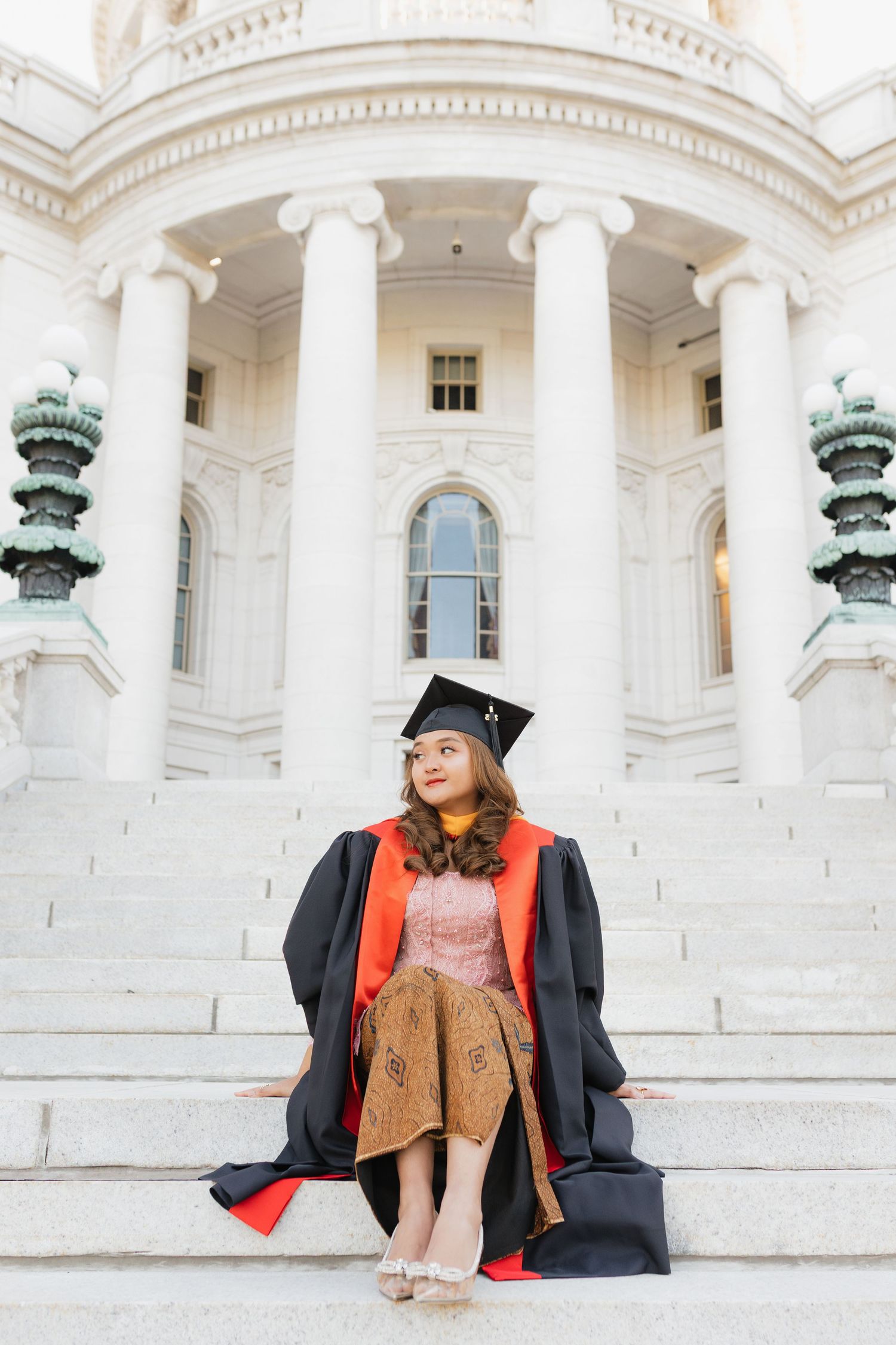 A UW Madison graduate in a black gown poses with their graduation cap at the Wisconsin State Capitol Building.