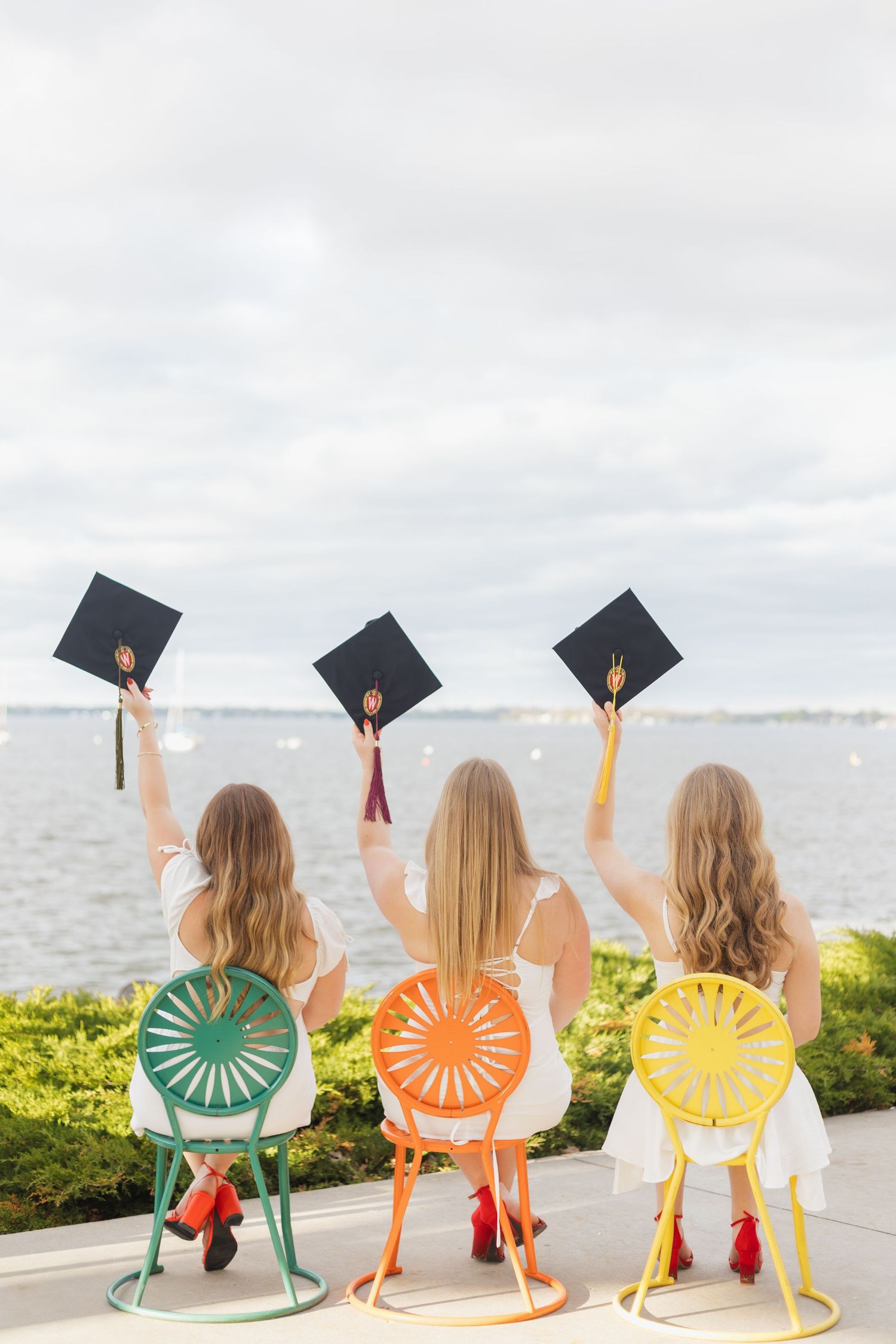 UW Madison graduates in colorful chairs raise their caps by the water on graduation day while sitting in yellow, orange and green seats.