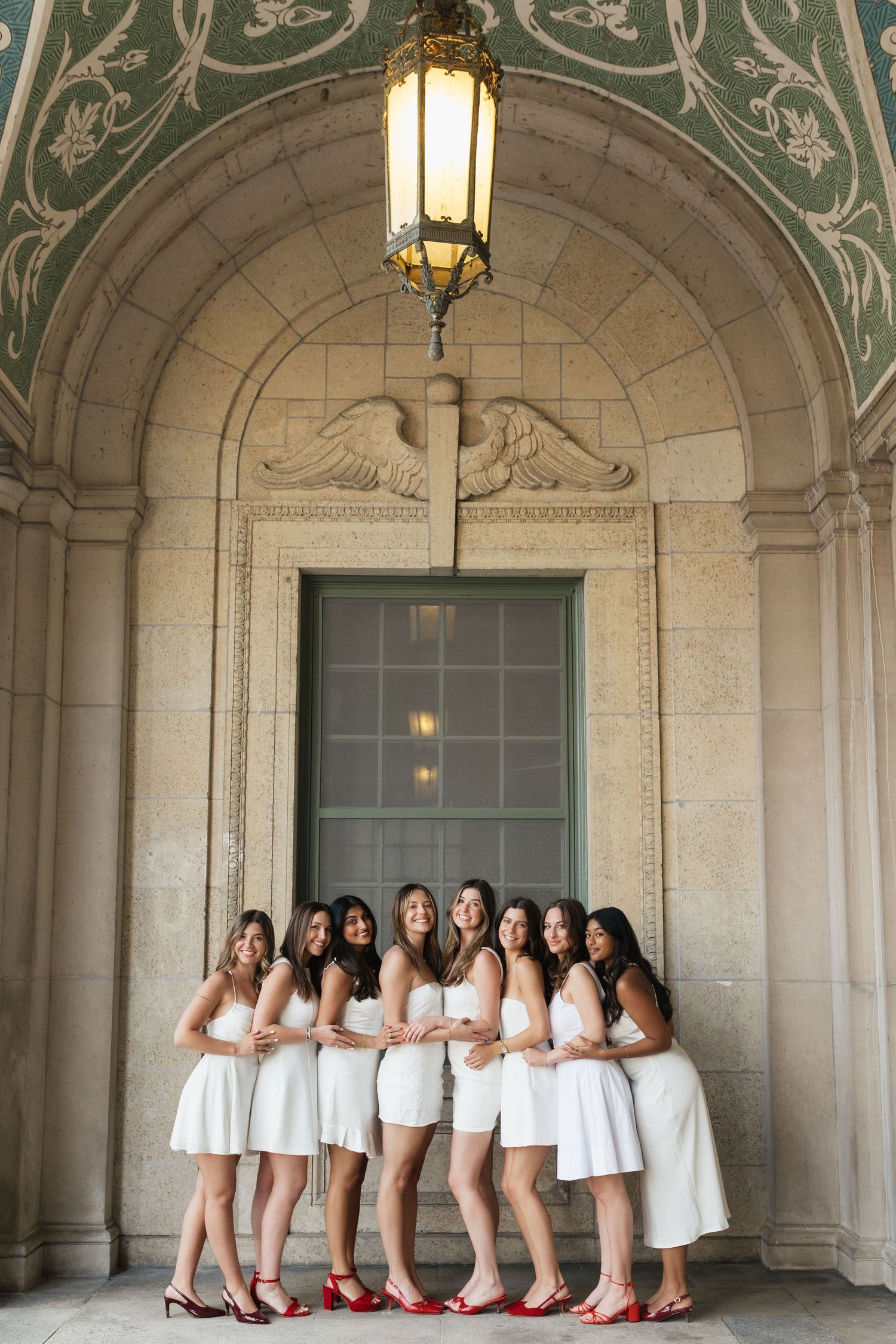 Group photo of women in white dresses posing under ornate archways with red shoes.