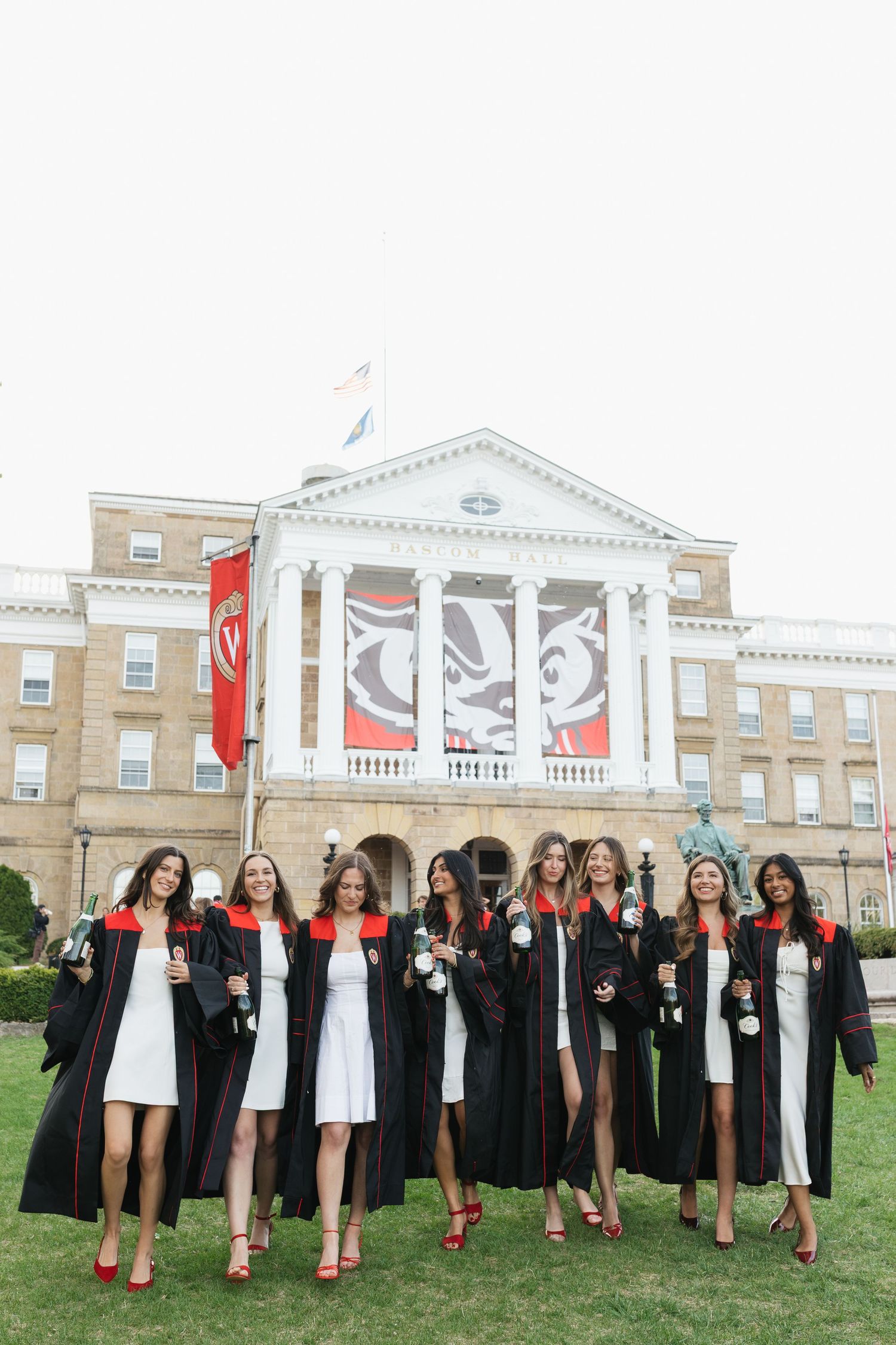 Graduates in black gowns and white dresses pose in front of a stately university building with red banners.