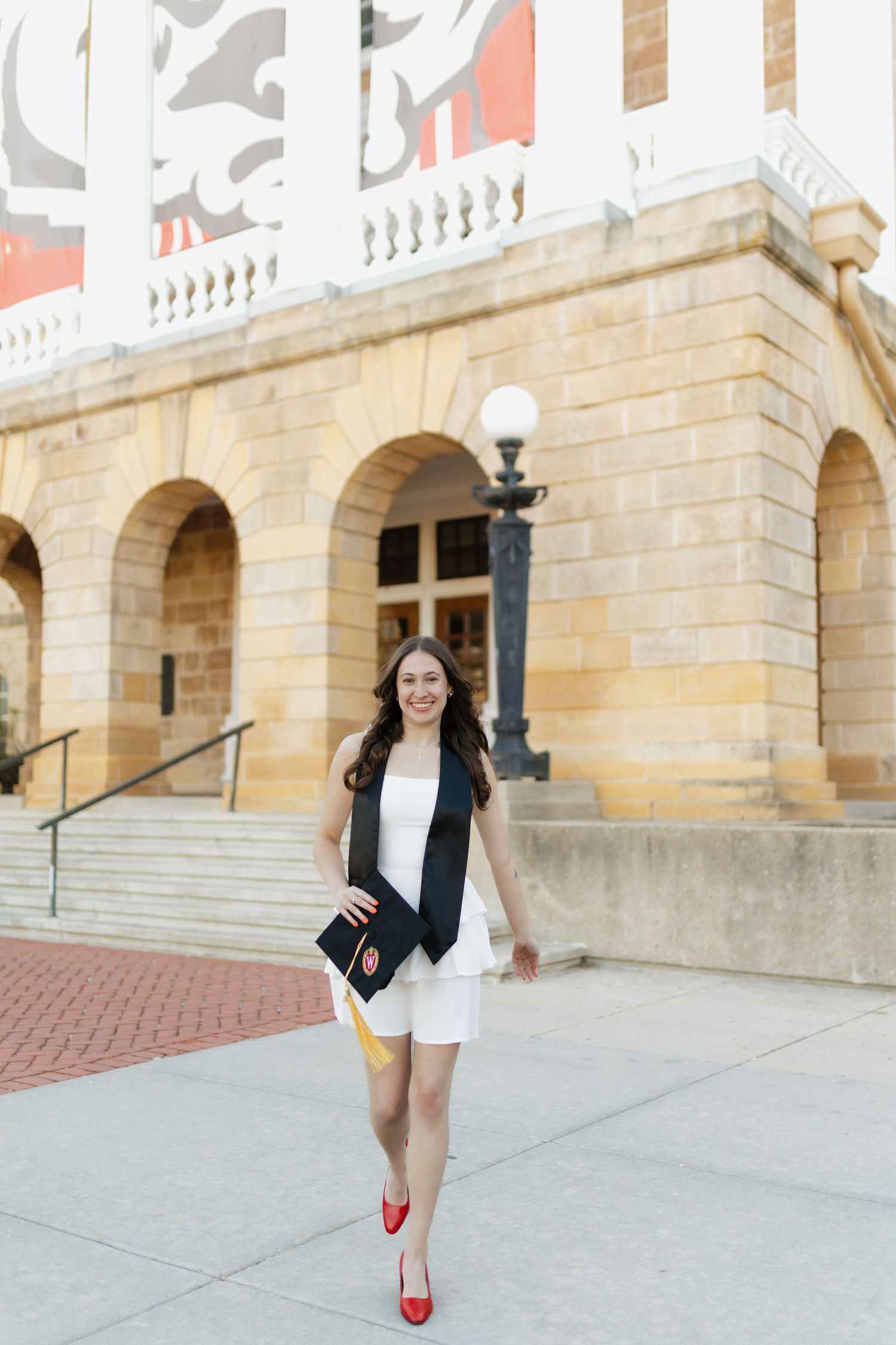 Graduate in black gown and red heels poses confidently on campus steps.