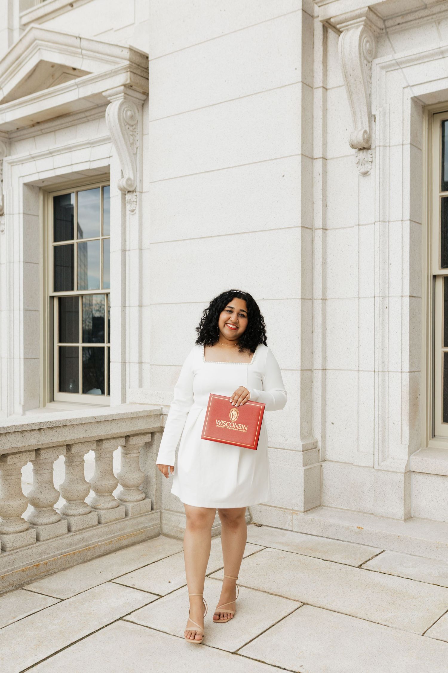 Graduate in white dress poses with red gift box against white stone architecture.