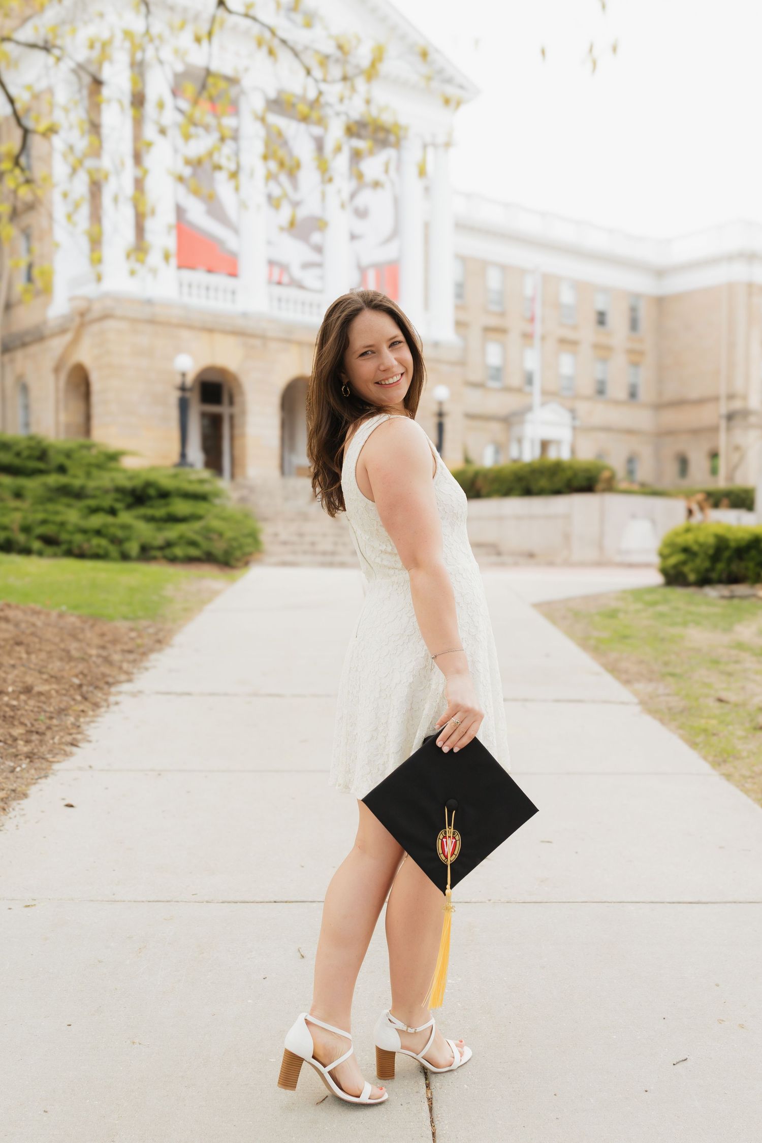 UW Madison graduate in a white dress holding a black cap and tassel along a stone building pathway.