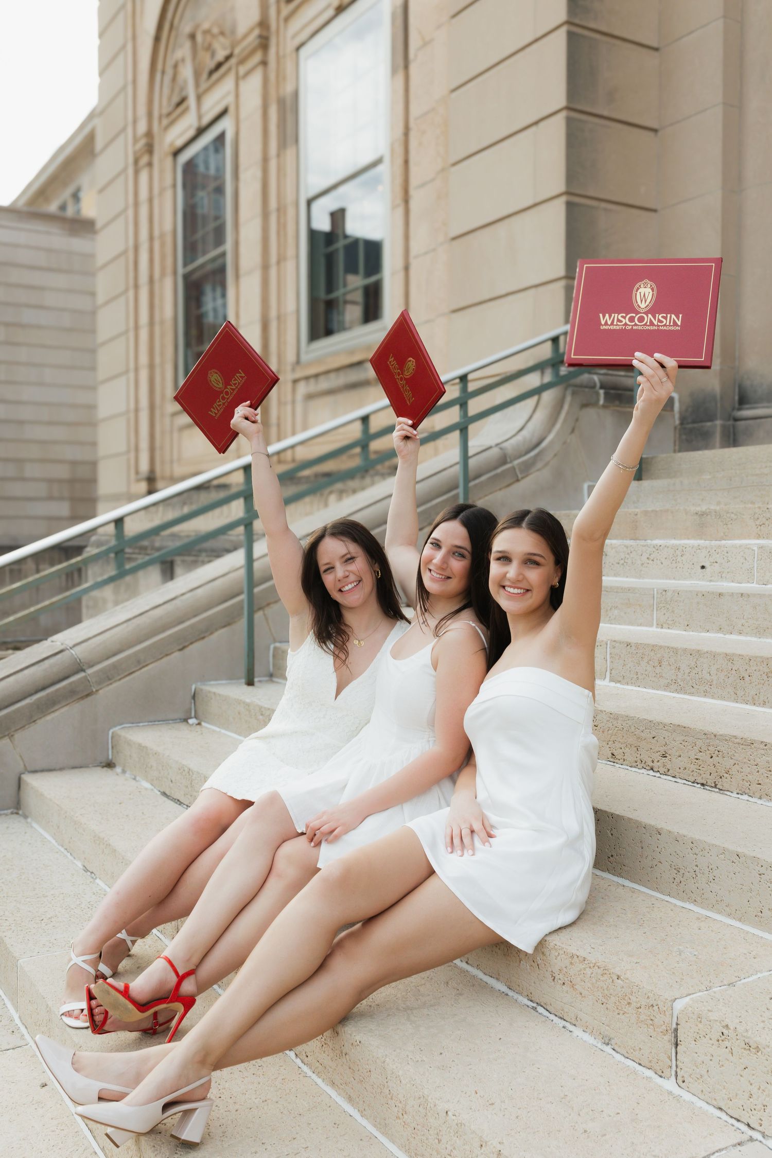 Three graduates in white dresses hold up red diplomas while sitting on stone steps.