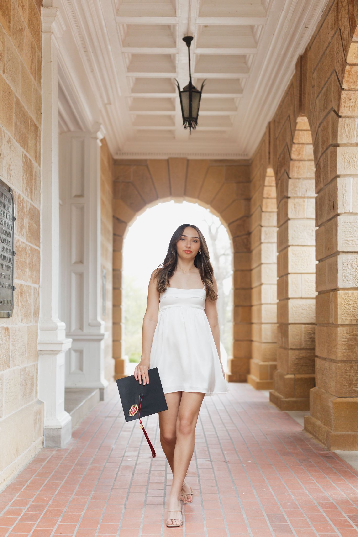 Graduate in white dress walks through columned university hallway.