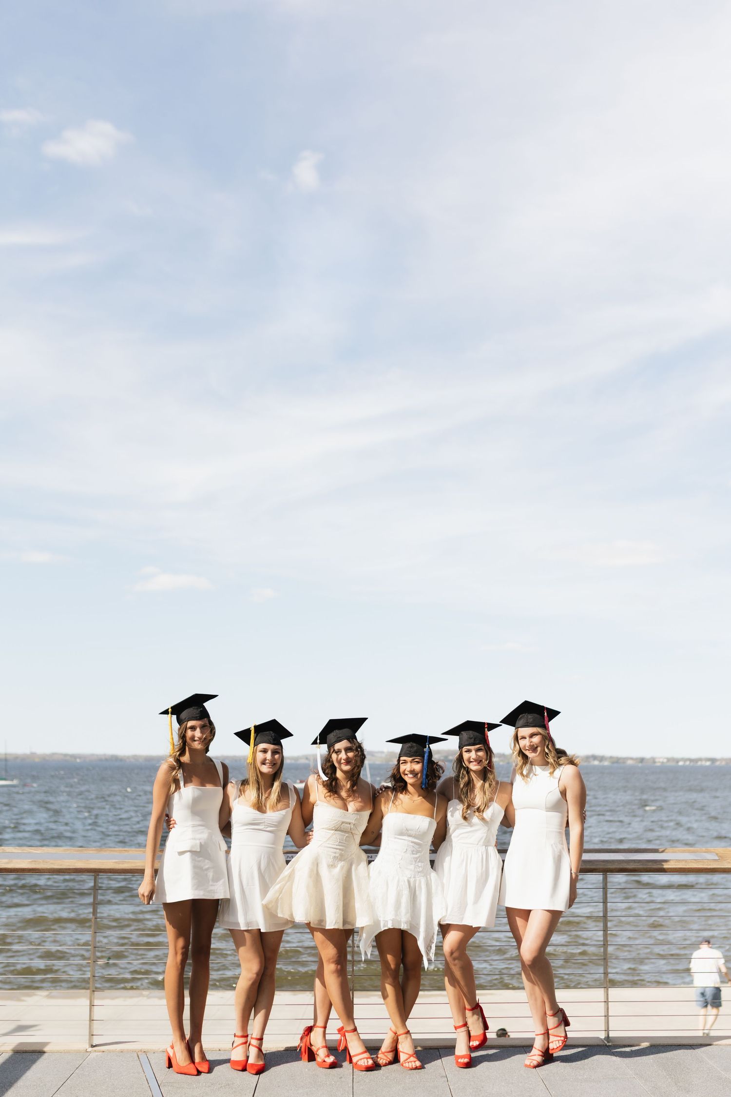 Graduates in white dresses and graduation caps pose by waterfront with red shoes.
