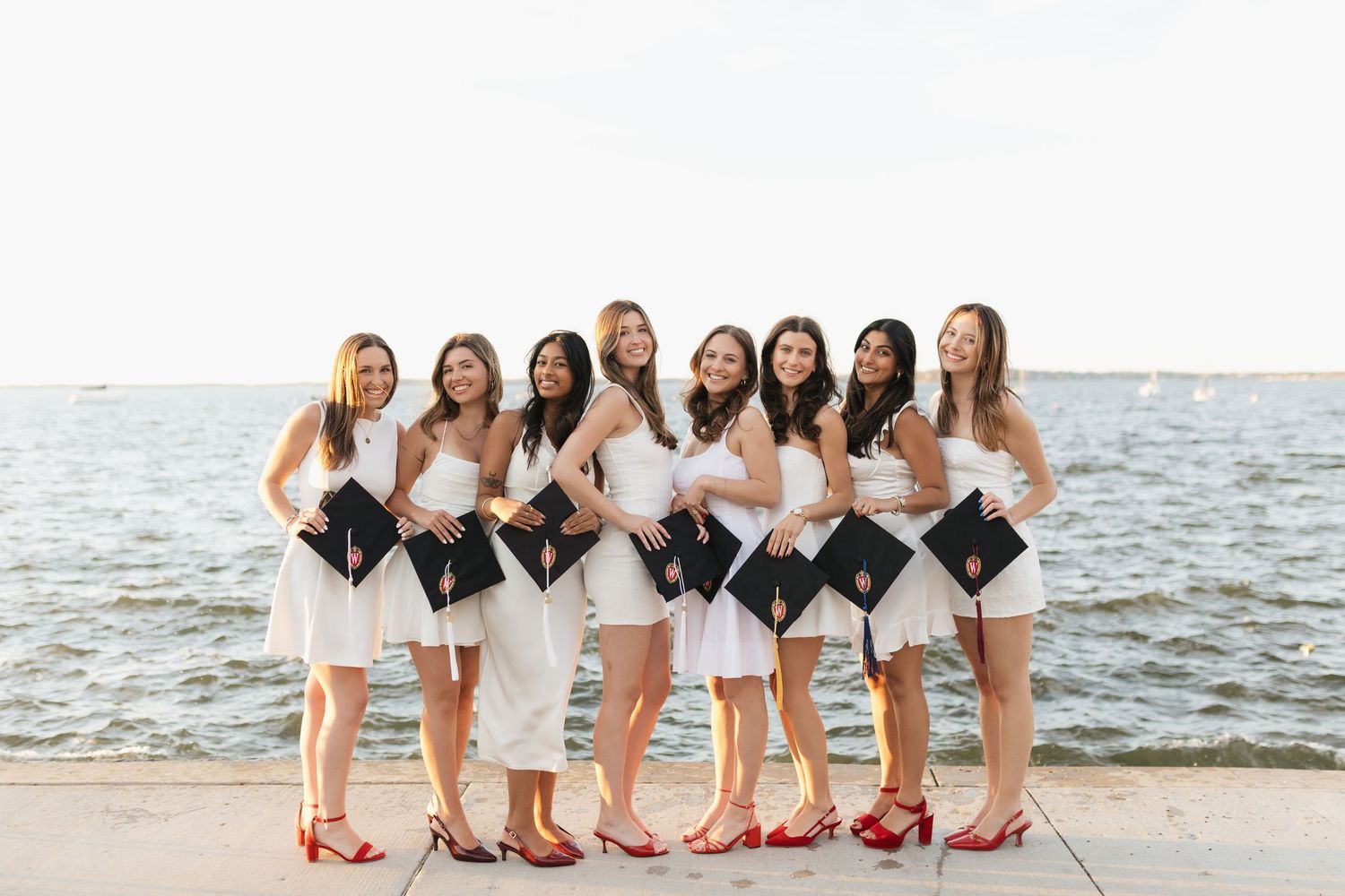 Row of graduates in white dresses hold diplomas while posing by waterfront.