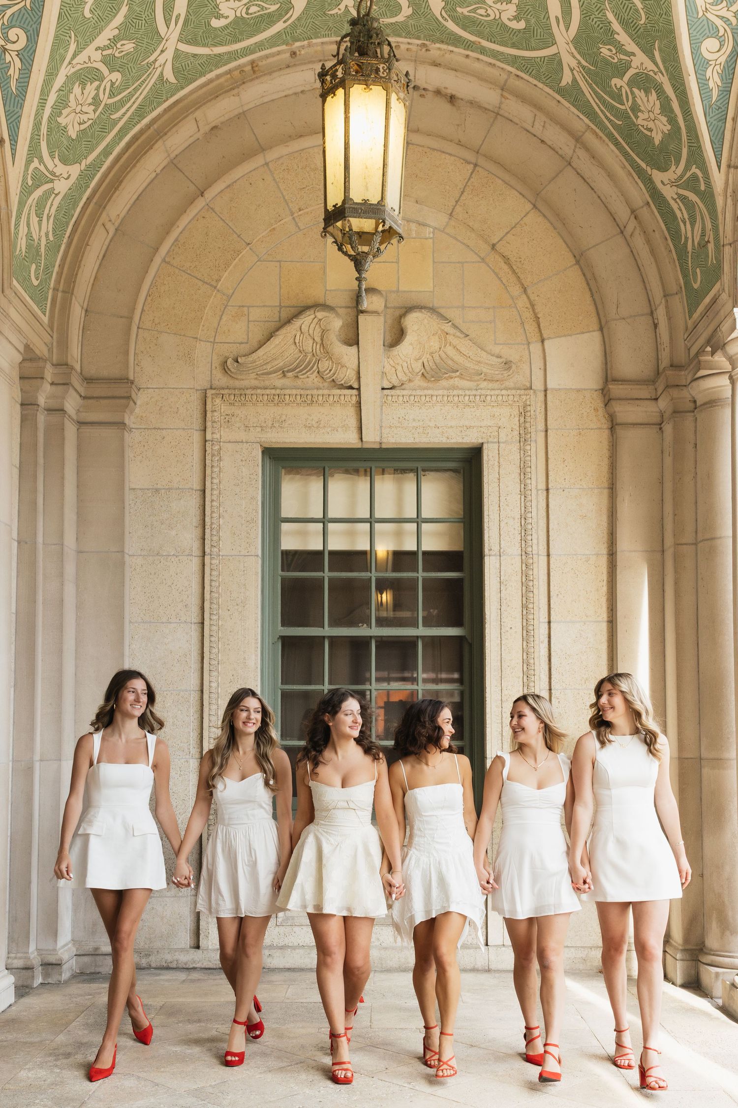 Graduates in white dresses and red heels pose in line under ornate architecture.