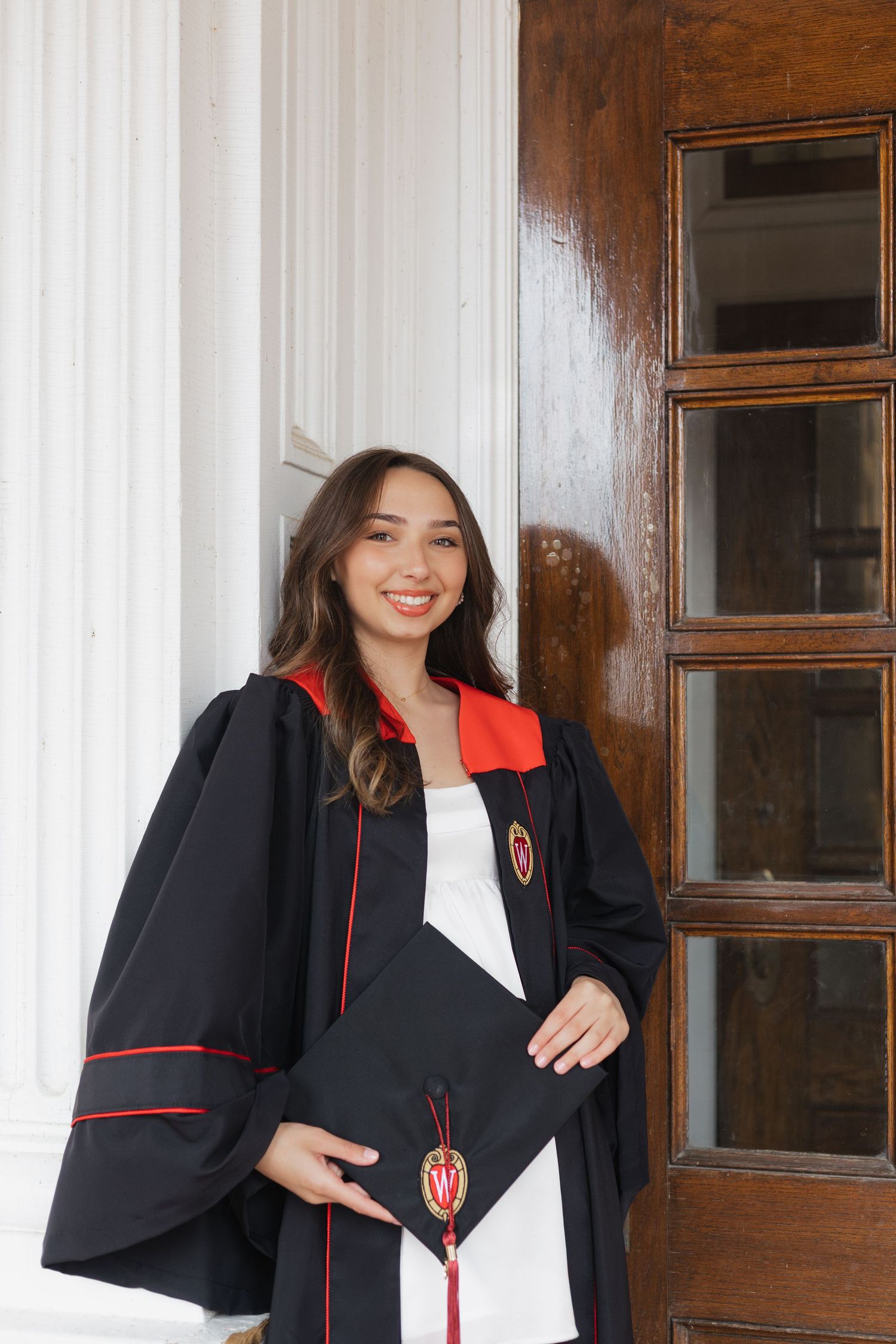 Graduate posing in black and red academic regalia against wooden door backdrop.