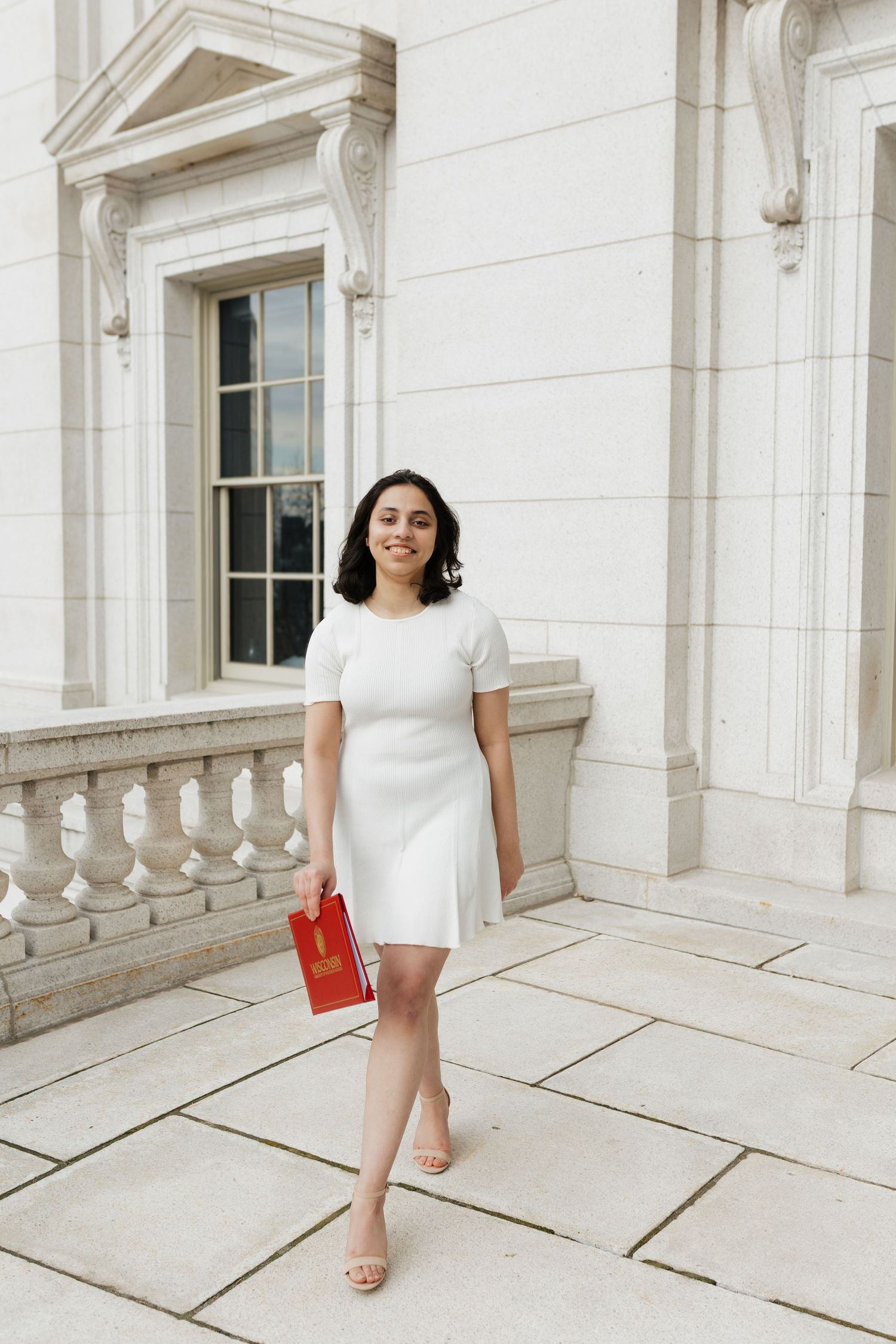 Graduate in white dress poses against classic white stone building columns.