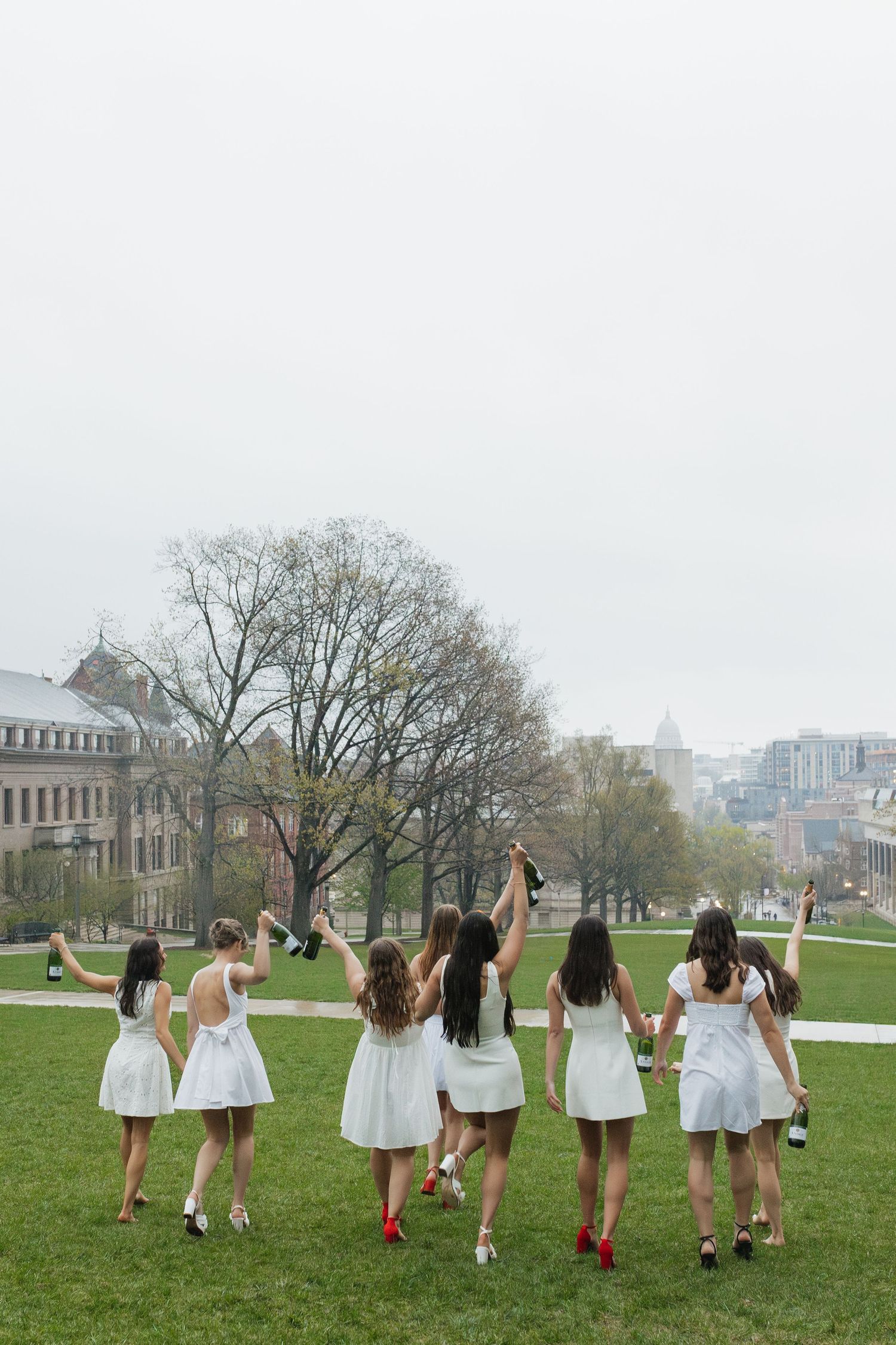 Graduates in white dresses walk across campus lawn holding diplomas.